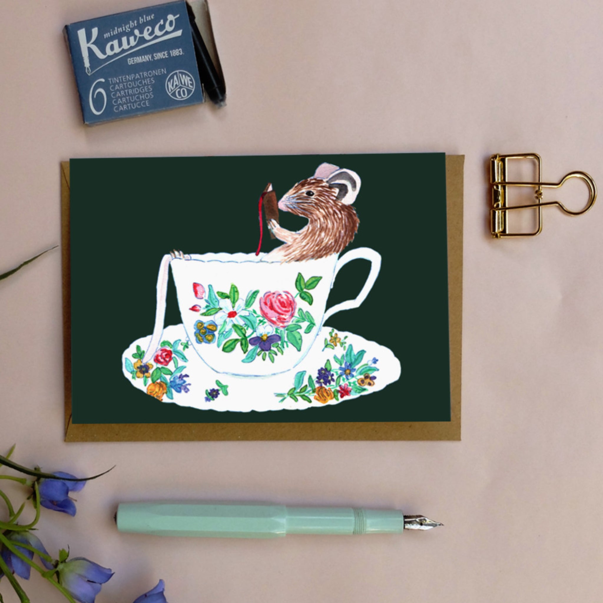 Illustration einer Maus mit Hut und Buch in einer geblümten Tasse, daneben Patronen, Clips und blaue Blumen.