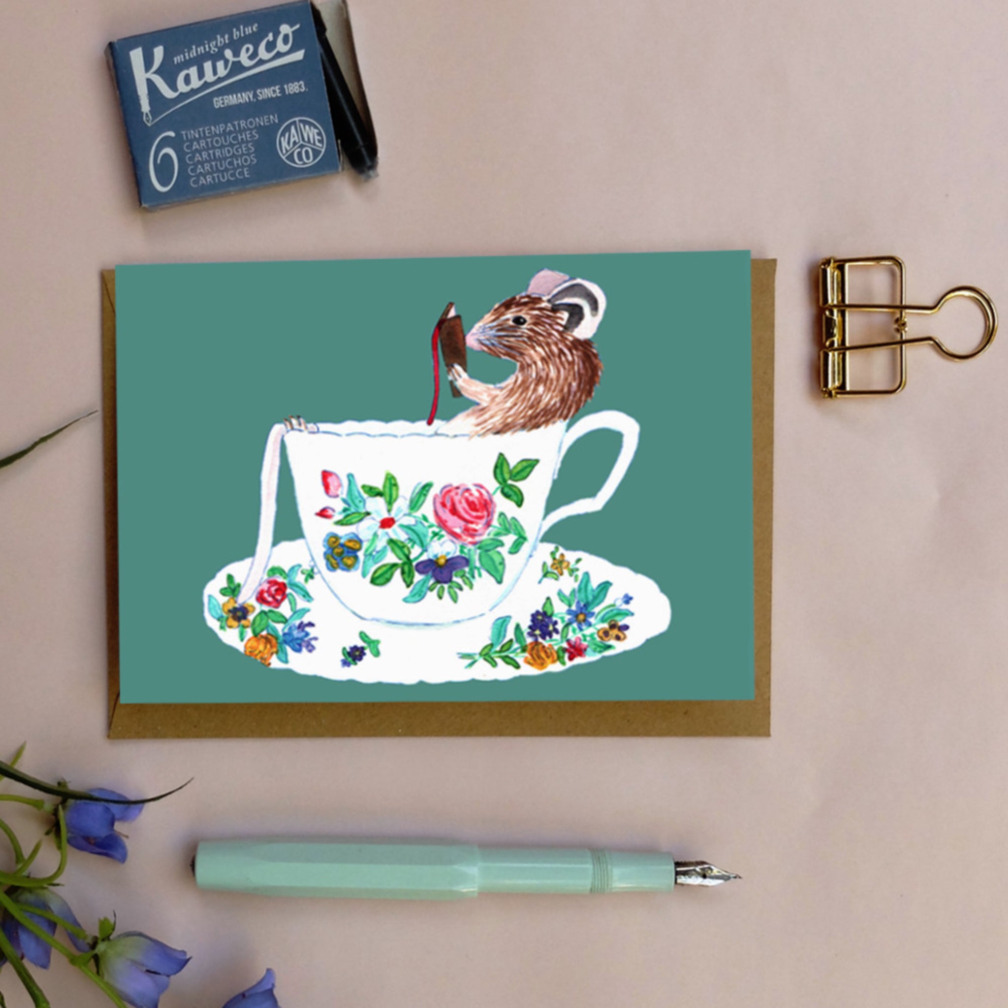 Eine Karte mit einer Zeichnung einer Maus, die in einer Teetasse mit Blumenmuster sitzt, umgeben von Schreibutensilien.