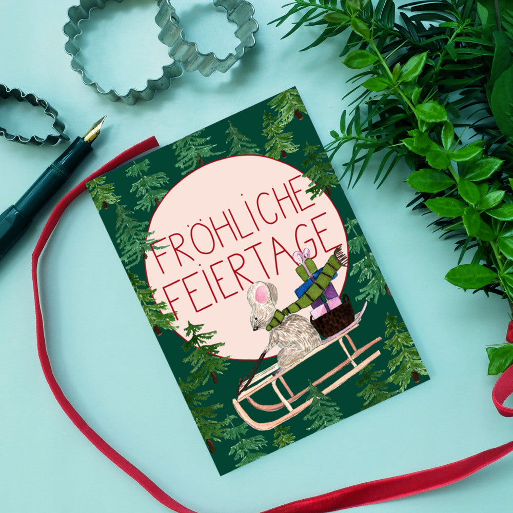Weihnachtskarte mit einer Maus auf einem Schlitten, umgeben von Tannenbäumen und dem Text "Fröhliche Feiertage".