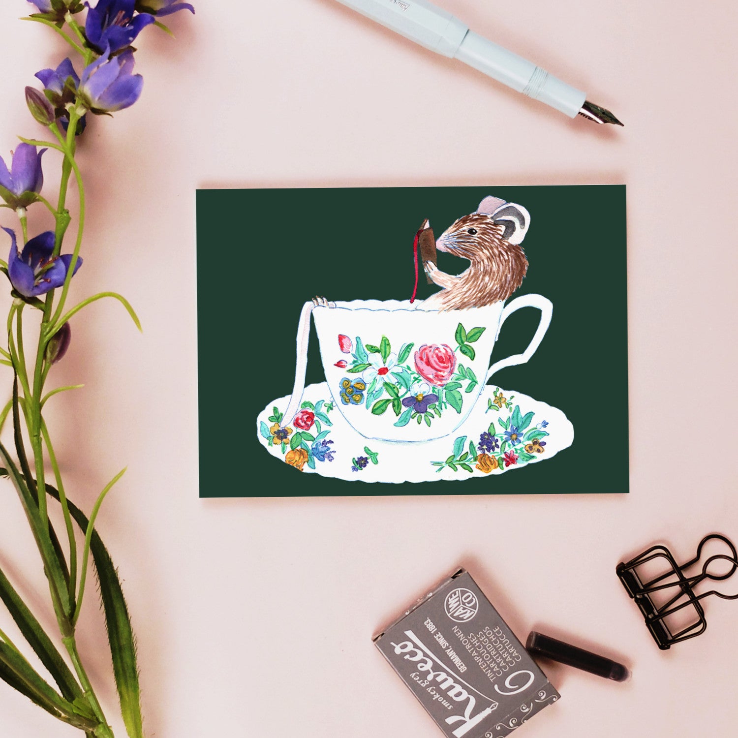 Eine Postkarte mit einer Illustration einer Maus in einer blumengeschmückten Teetasse, umgeben von Schreibutensilien.