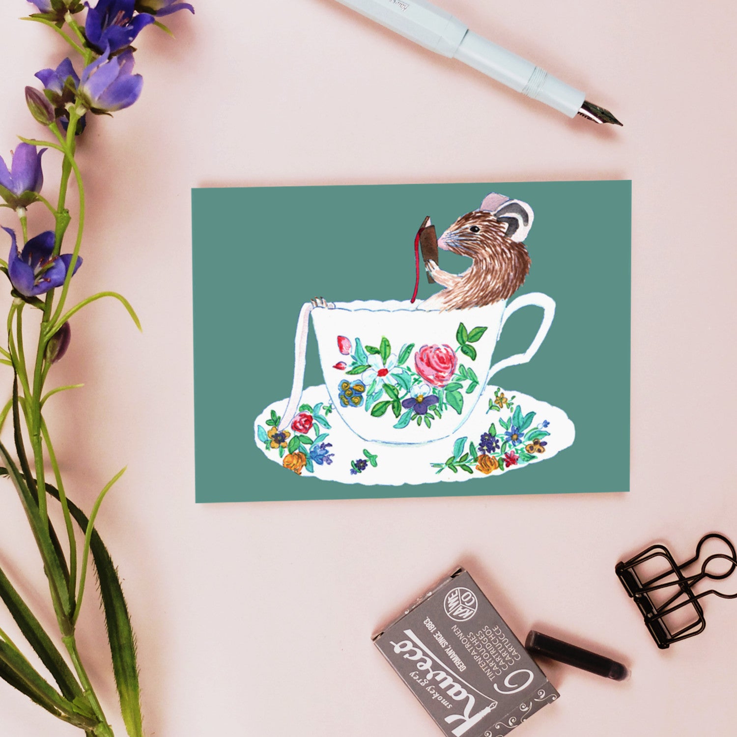Eine Postkarte mit der Abbildung einer Maus in einer geblümten Teetasse liegt auf einer rosafarbenen Fläche neben einem blauen Stift, einem grauen Radiergummi, einer Büroklammer und einem Zweig mit lila Blumen.