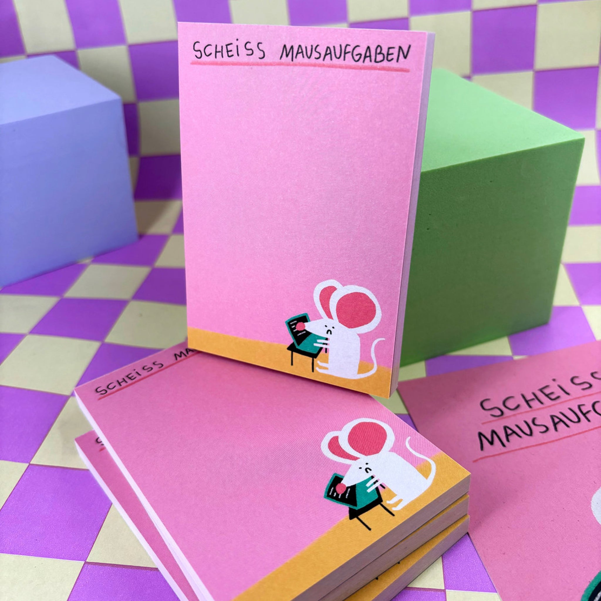 Notizblöcke mit der Aufschrift "Scheiss Mausaufgaben" und einer Zeichnung einer Maus am Computer auf rosa Hintergrund.