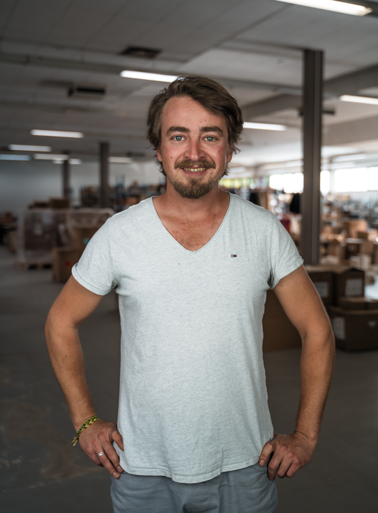 Ein Mann in einem weißen T-Shirt lächelt in die Kamera, während er in einer Lagerhalle mit Kartons steht.
