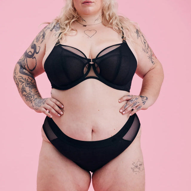 Eine übergroße Person mit langen blonden Haaren posiert selbstbewusst vor einem rosafarbenen Hintergrund und trägt einen passenden schwarzen BH und ein Unterwäsche-Set. Sie hat sichtbare Tattoos an Armen, Brust und Beinen.