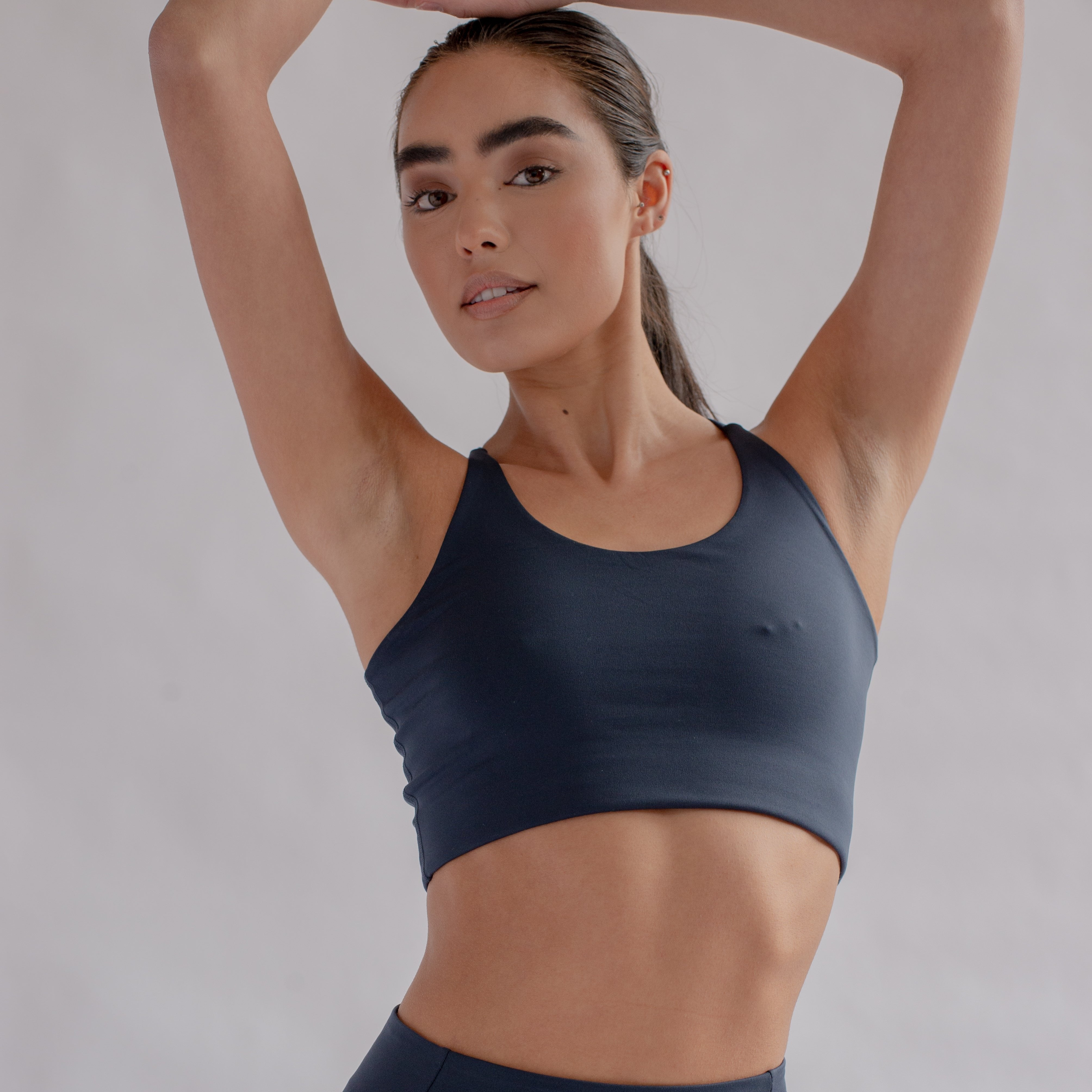 Frau in einem schwarzen Sport-BH hebt die Arme über den Kopf und lächelt sanft in die Kamera.