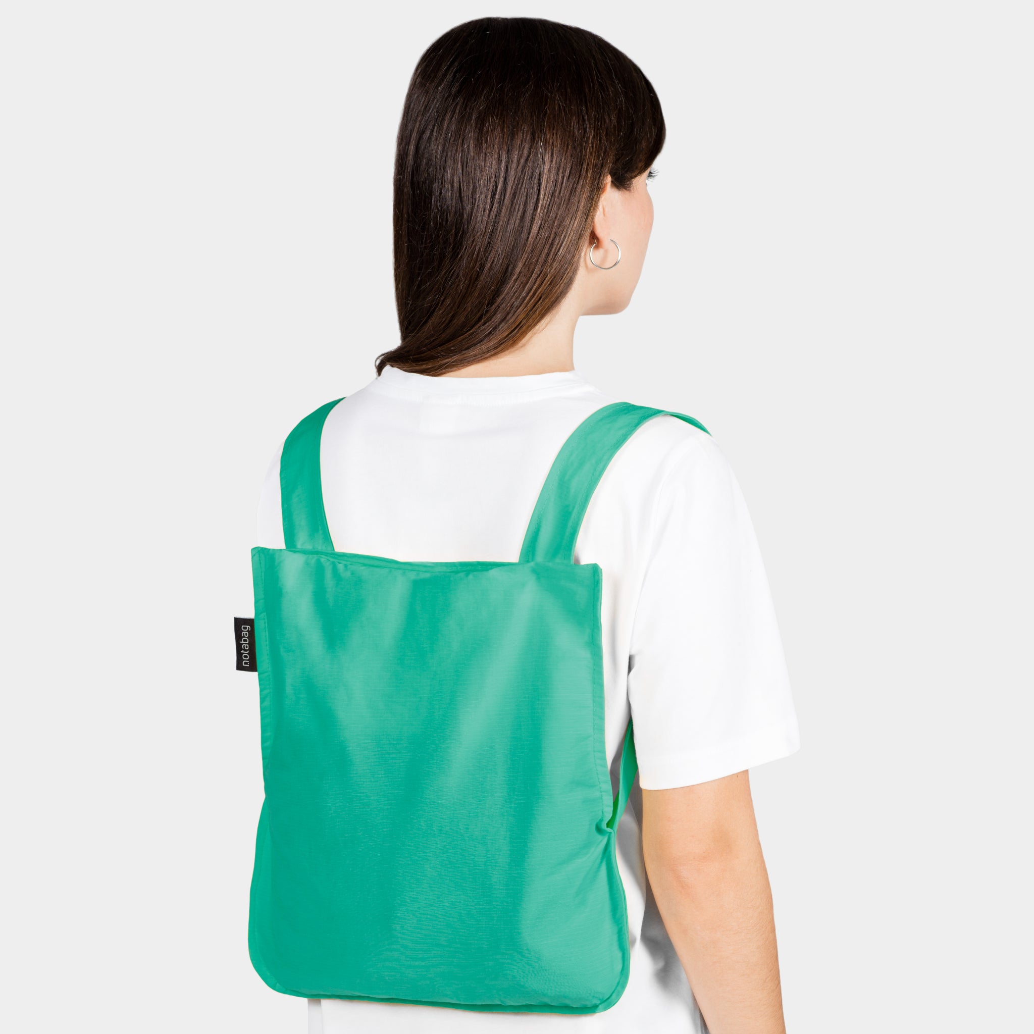 Person mit langen Haaren trägt einen grünen Rucksack und ein weißes T-Shirt, von hinten gesehen, auf grauem Hintergrund.