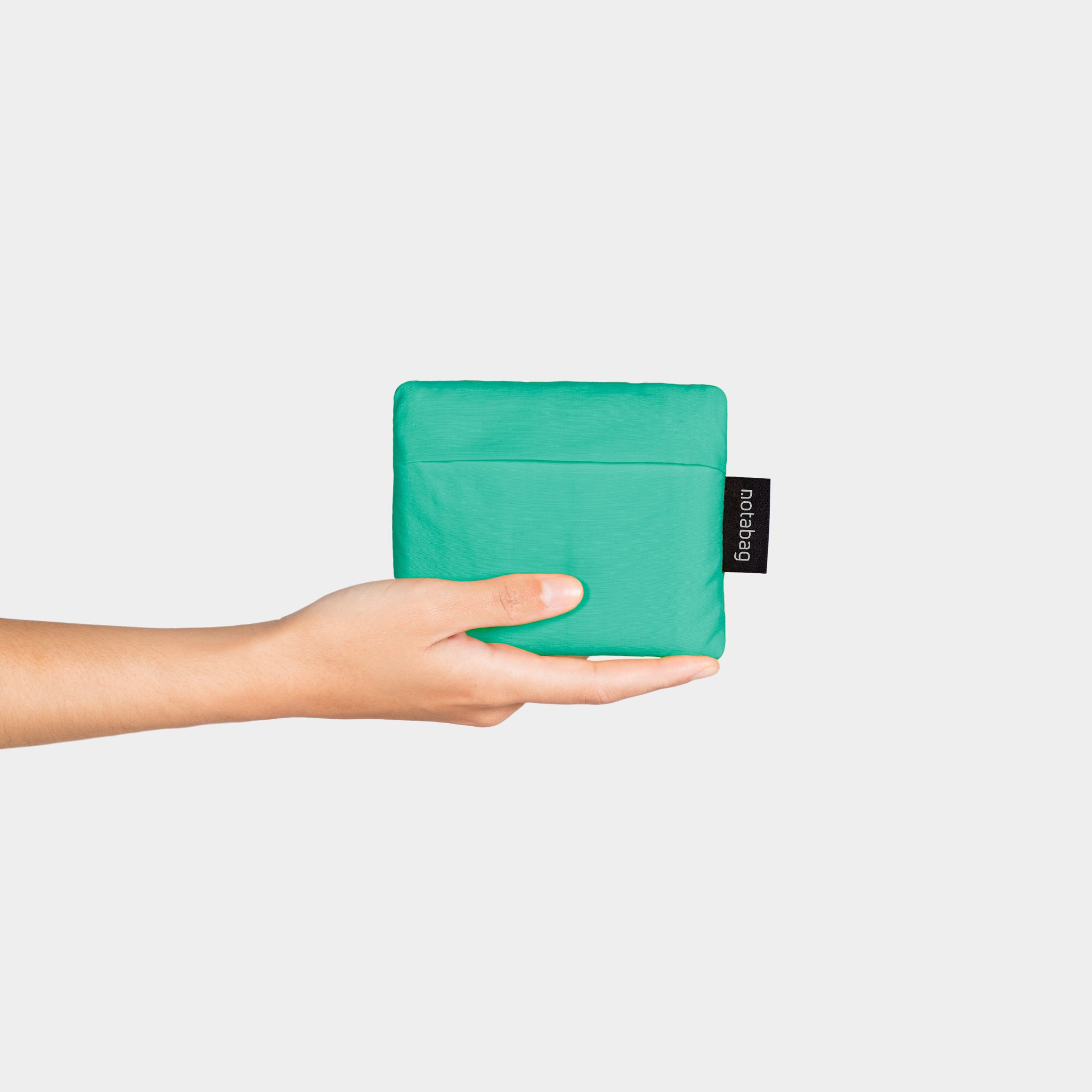 Hand hält eine grüne, gefaltete Tasche mit dem Etikett "notabag" vor einem weißen Hintergrund.