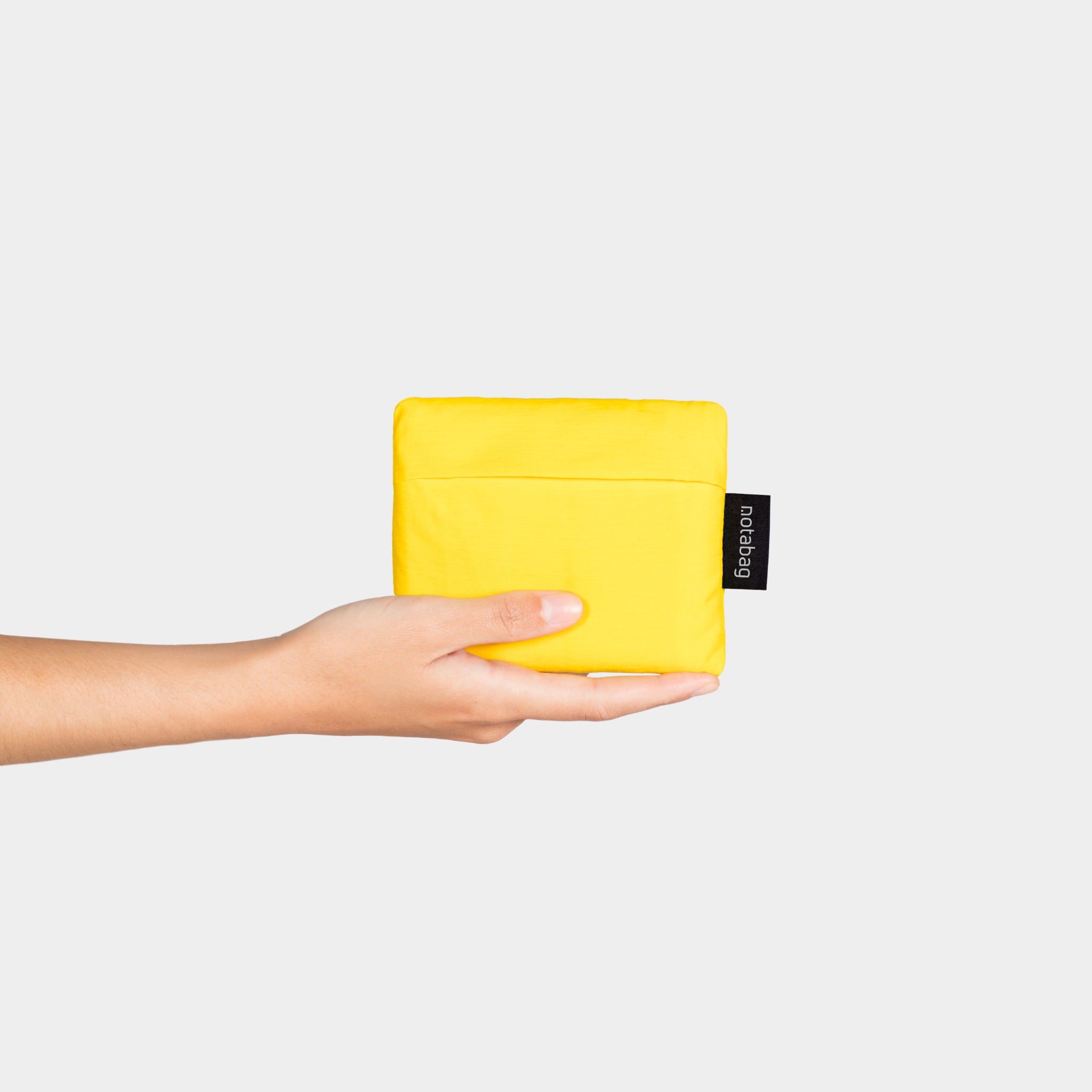 Eine Hand hält eine gelbe, quadratische Tasche mit einem kleinen schwarzen Etikett vor einem weißen Hintergrund.