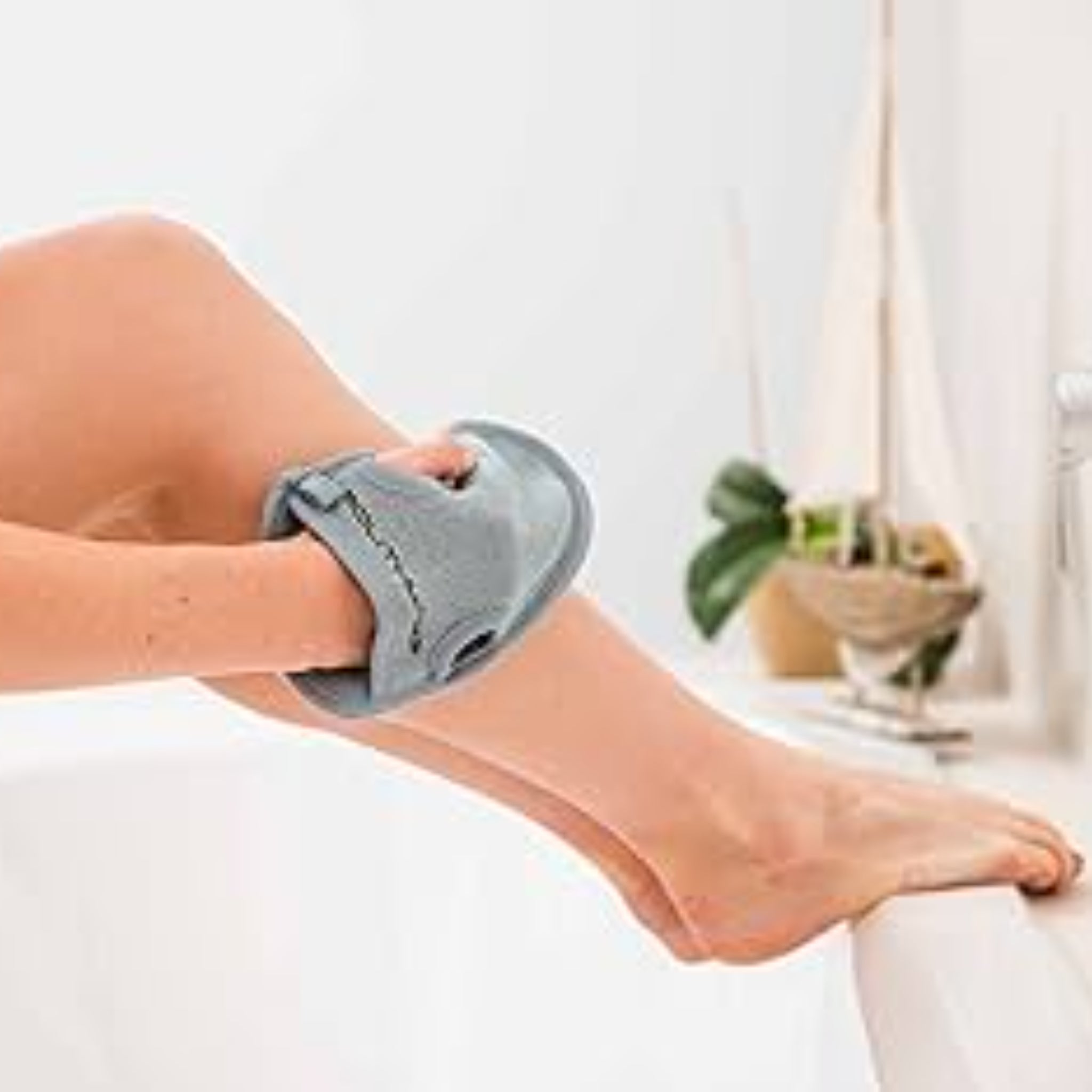 Eine Person wäscht ihr Bein mit einem grauen Waschhandschuh in der Badewanne, im Hintergrund sind Pflanzen sichtbar.