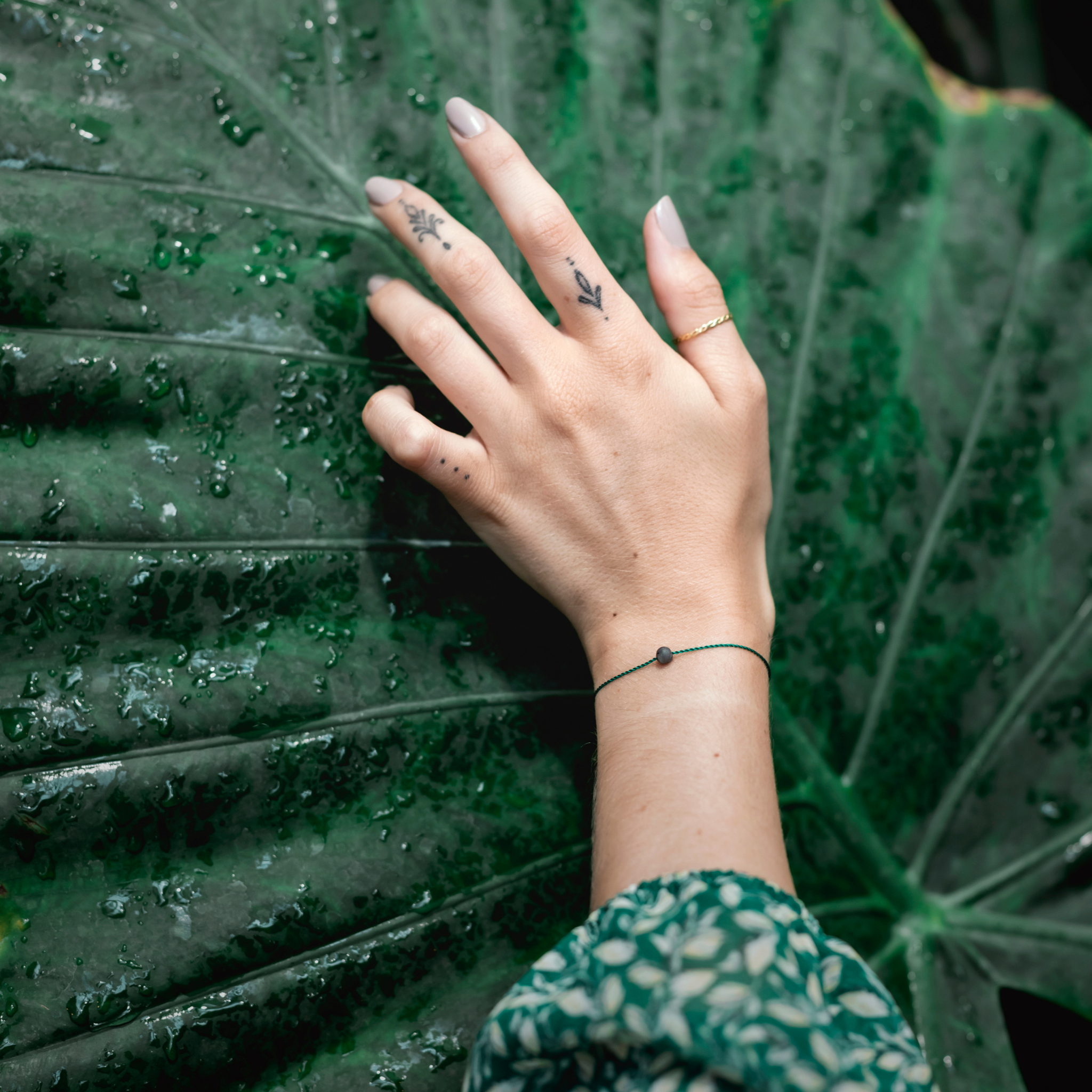 Hand mit Tattoos und Armband berührt ein großes, nasses Blatt.