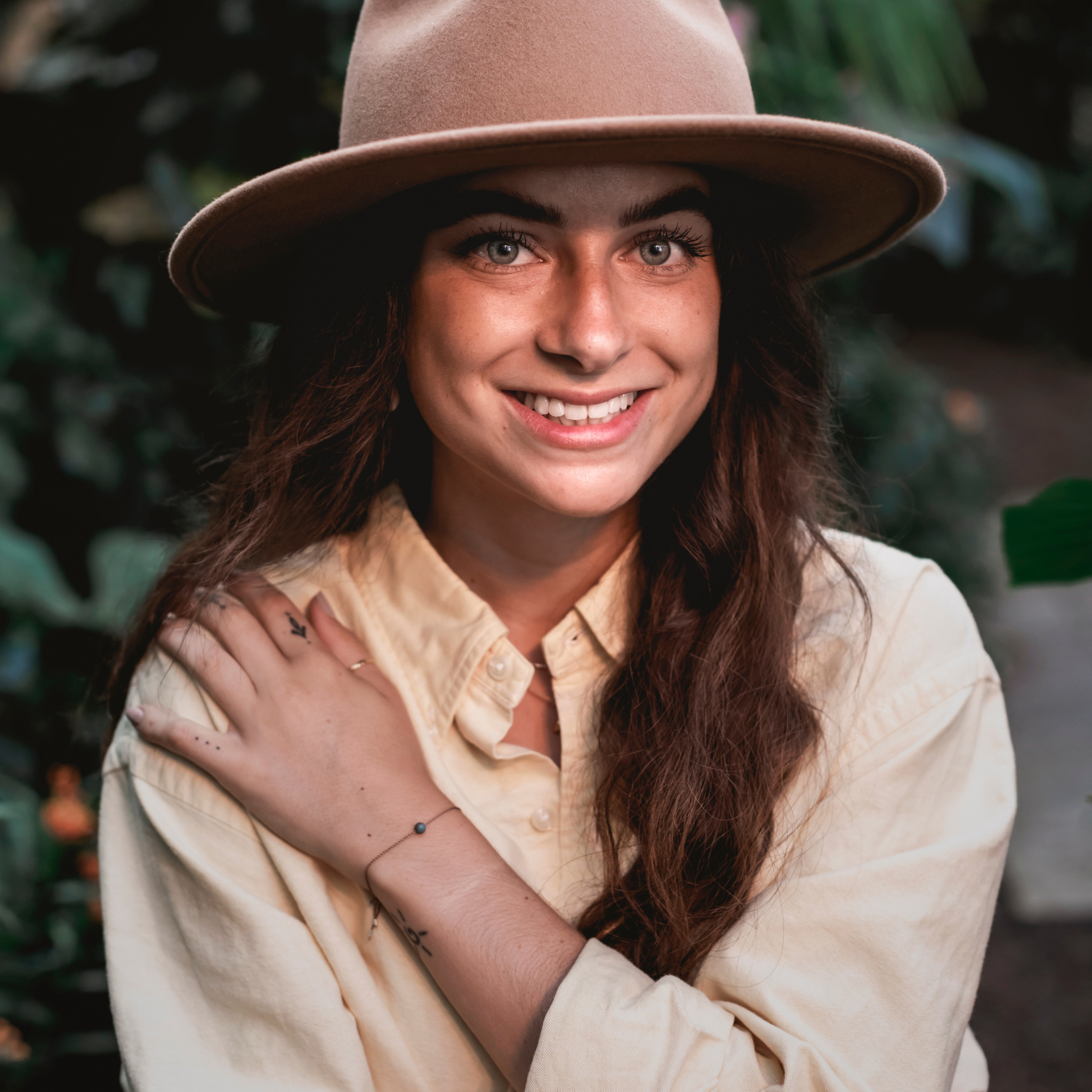 Frau mit braunem Hut lächelt in die Kamera, trägt ein helles Hemd und hat den Arm leicht angehoben. Hintergrund ist grün.