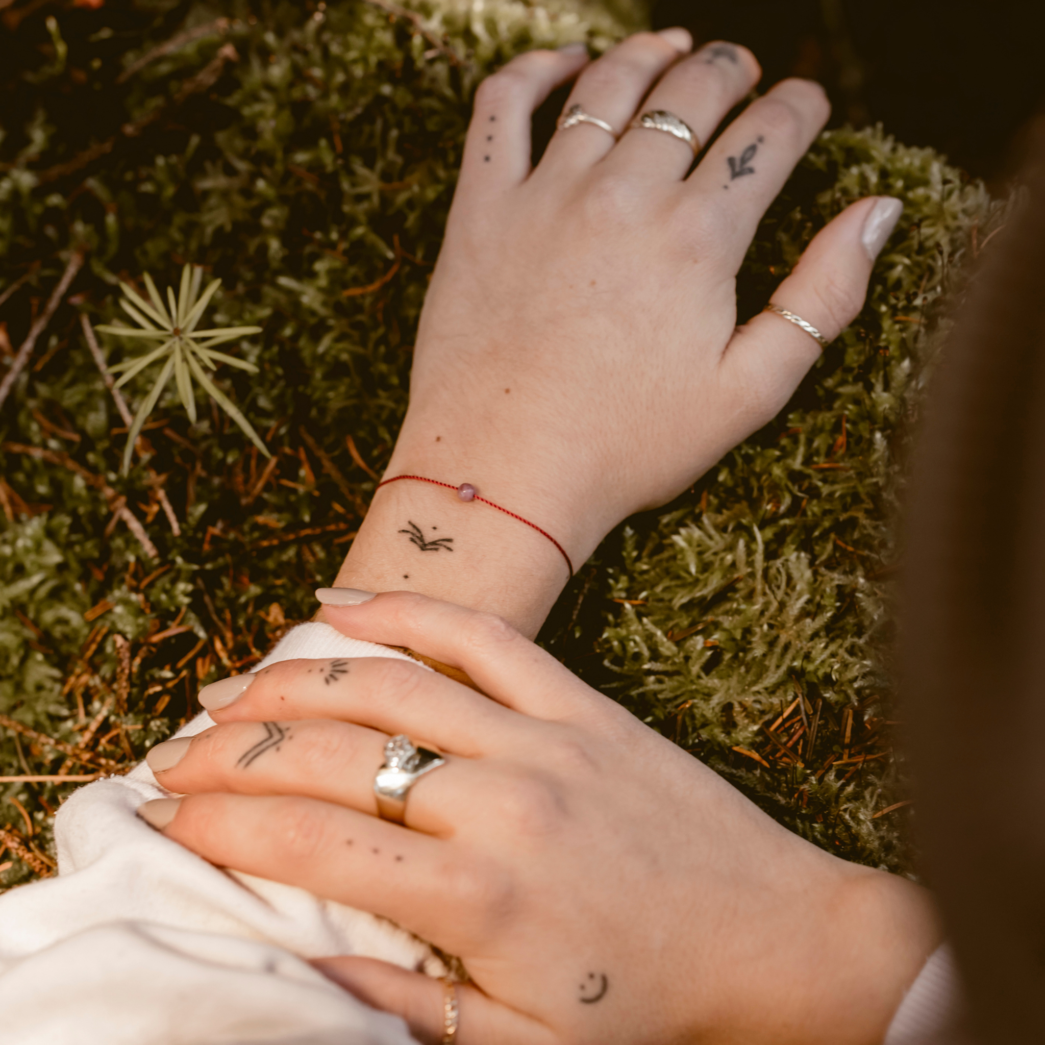 Hände mit Tattoos auf Moos; ein rotes Armband und mehrere Ringe sind sichtbar.