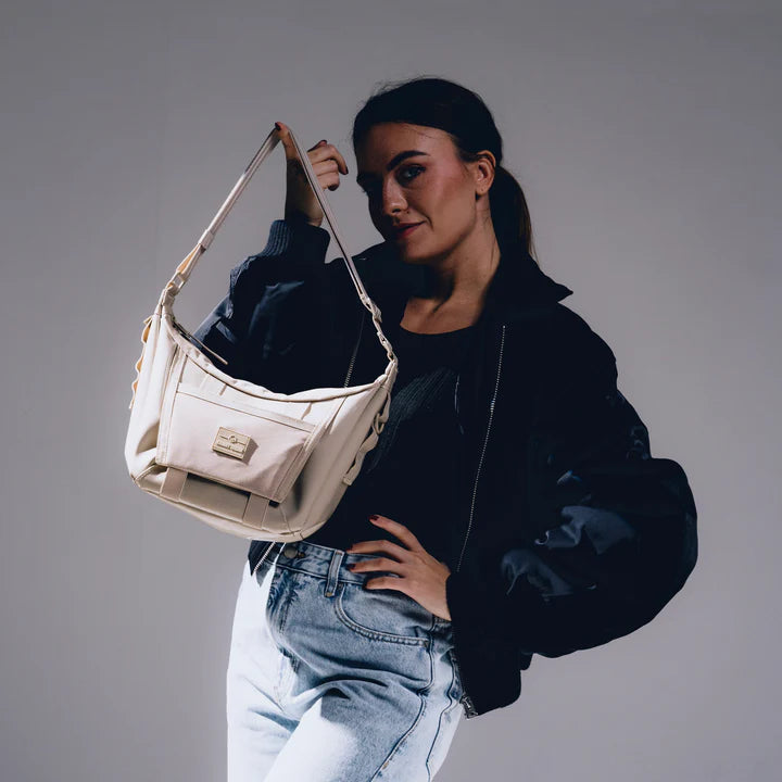 Eine Frau in schwarzer Jacke und hellblauen Jeans posiert selbstbewusst und hält eine beigefarbene Umhängetasche mit einem rechteckigen Logo vor einem neutralen Hintergrund in die Kamera.