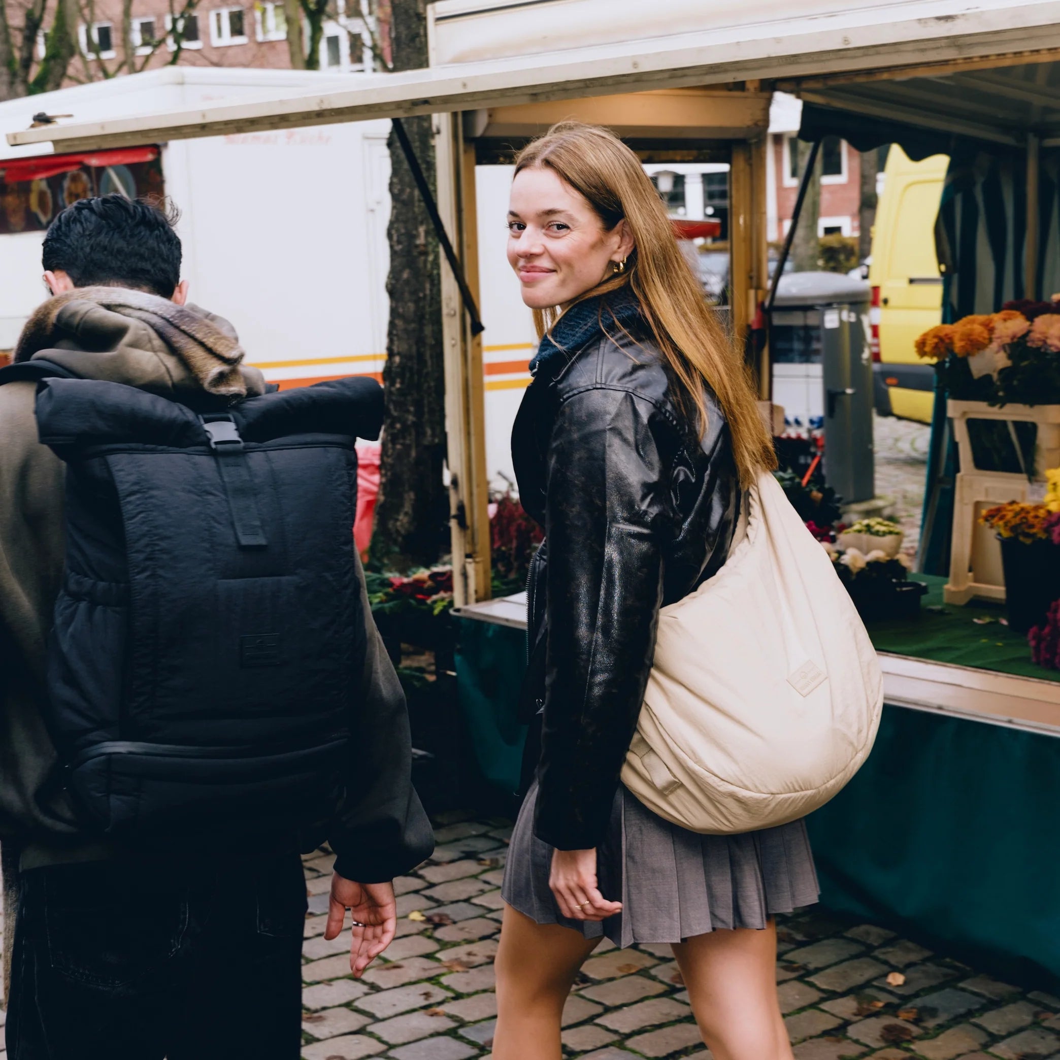 Eine Frau in einer schwarzen Lederjacke und einem grauen Rock lächelt in die Kamera, während sie mit einer großen beigen Tasche durch einen Markt geht. Ein Mann mit einem schwarzen Rucksack geht neben ihr her. Blumen werden an einem Marktstand ausgestellt.