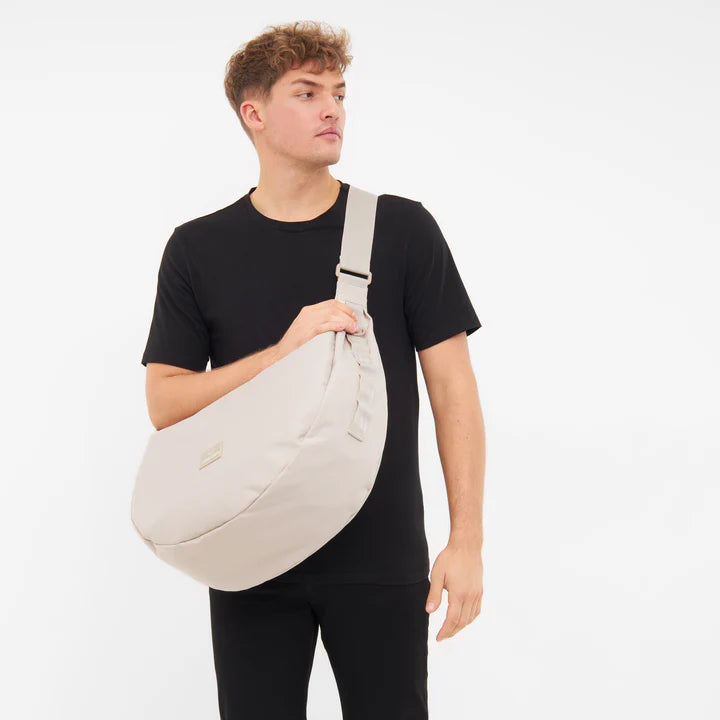 Eine Person, die ein schwarzes T-Shirt und eine schwarze Hose trägt, steht vor einem schlichten weißen Hintergrund und trägt eine große beigefarbene Umhängetasche quer über den Körper.