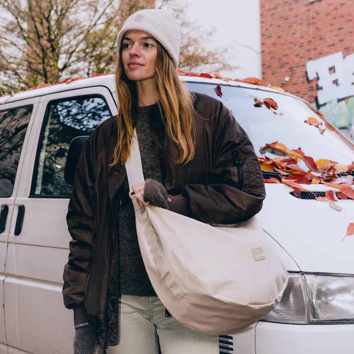 Eine Frau in einer braunen Jacke und einer beigen Mütze steht vor einem weißen Lieferwagen mit Herbstblättern auf der Motorhaube. Sie trägt eine große beigefarbene Umhängetasche und blickt zur Seite, wo Bäume und eine Backsteinmauer im Hintergrund zu sehen sind.