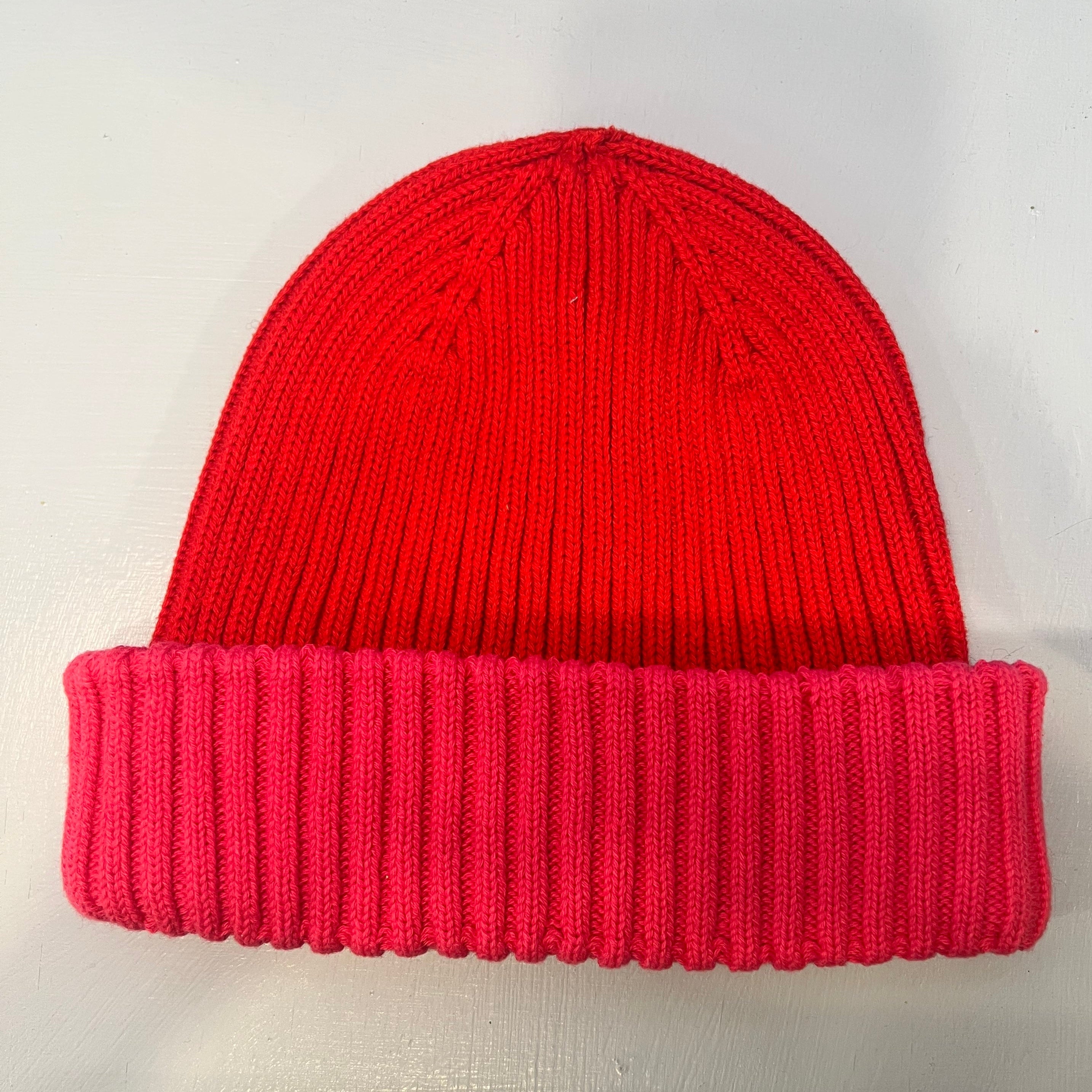 Eine gestrickte Beanie-Mütze mit leuchtend rotem Oberteil und gefaltetem, geripptem rosa Rand, dargestellt auf einer hellgrauen Fläche.