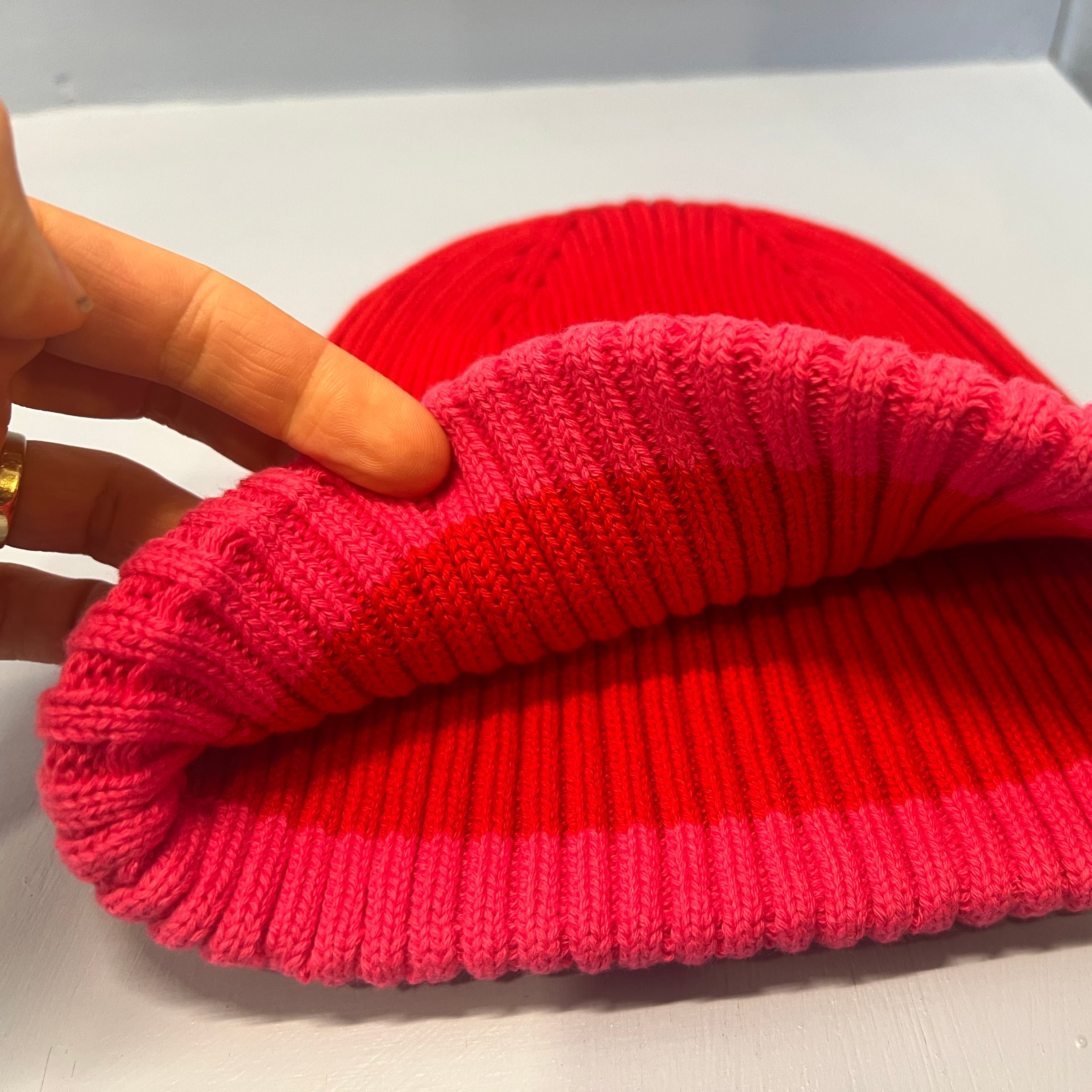 Eine Hand hebt den Rand einer Rippenstrickmütze an und enthüllt das leuchtend rosa Innere und das rote Äußere, beide aus demselben gerippten Material, auf einer hellgrauen Oberfläche.