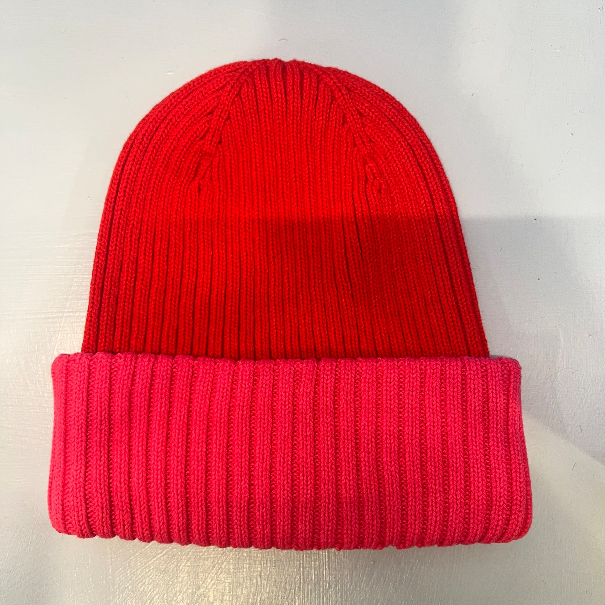Rippenstrickmütze mit rotem Scheitel und breiter, hellrosa gefalteter Krempe auf hellgrauem Untergrund.