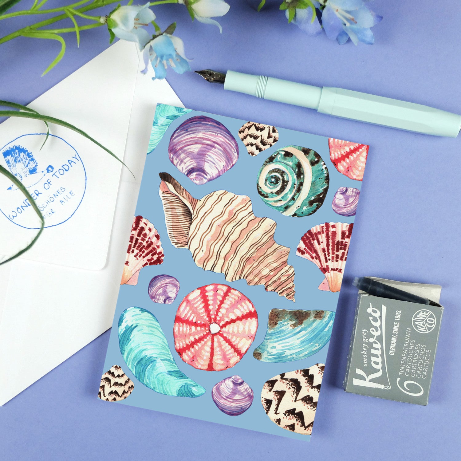 Karte mit bunten Muscheldesigns auf blauem Hintergrund, umgeben von einem Füllfederhalter, Tinte und Blumen.