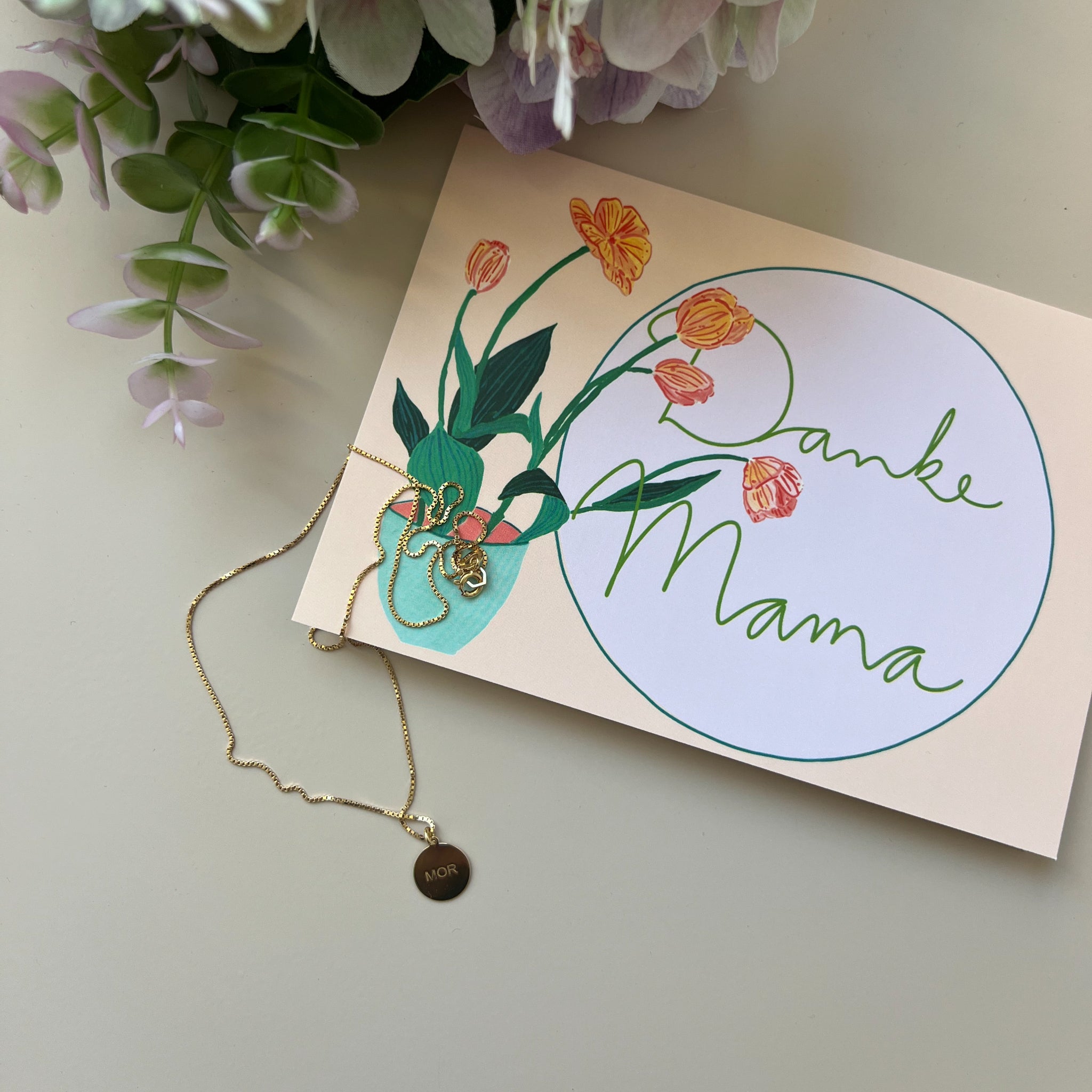 Grußkarte mit Aufschrift "Danke Mama", verziert mit Tulpenzeichnung, daneben goldene Halskette mit "MOR"-Anhänger.