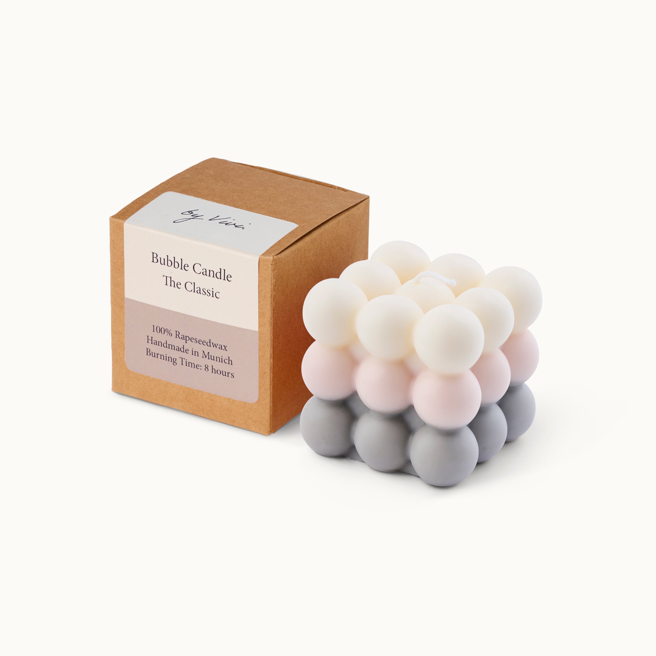 Eine würfelförmige Bubble Candle mit pastellrosa, weißen und grauen Kugeln steht neben einer braunen Schachtel mit der Aufschrift Bubble Candle The Classic. Auf der Schachtel steht, dass sie aus 100 % Bienenwachs handgefertigt ist.