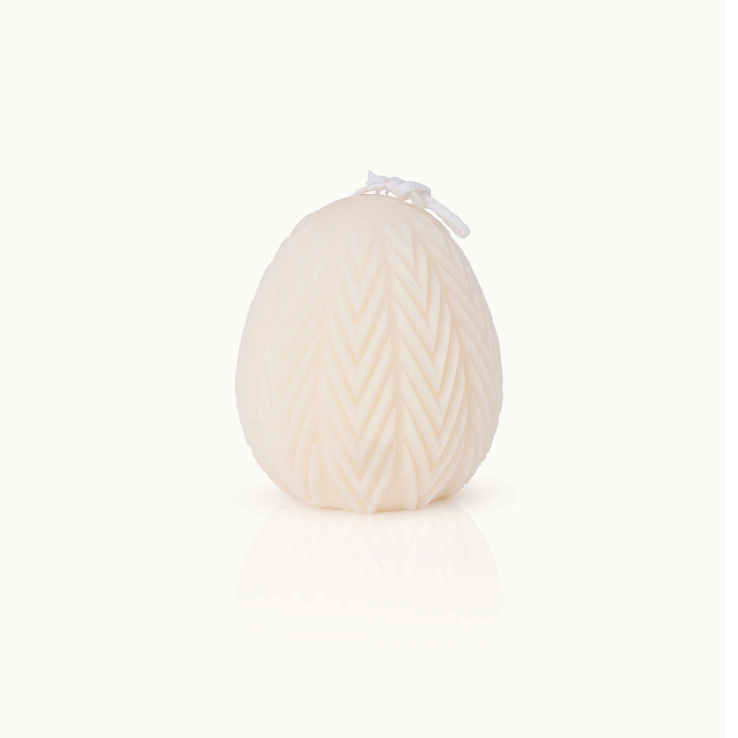 Eine weiße, eiförmige Kerze, in deren Oberfläche ein Chevron-Muster eingeritzt ist, vor einem schlichten weißen Hintergrund.