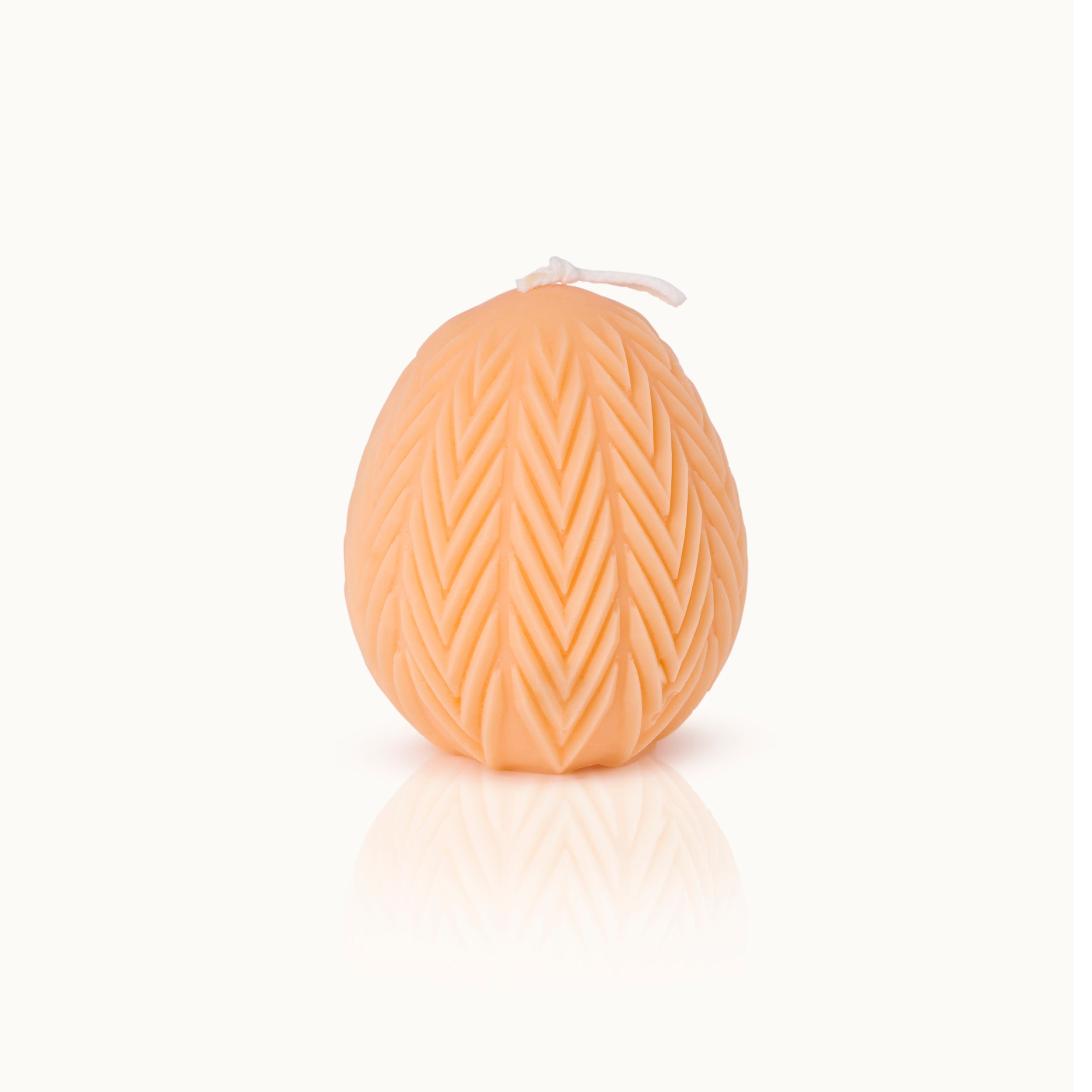 Eine pfirsichfarbene Kerze in Form eines Eies mit einem strukturierten Zickzackmuster und einem kurzen weißen Docht auf der Spitze vor einem schlichten weißen Hintergrund.