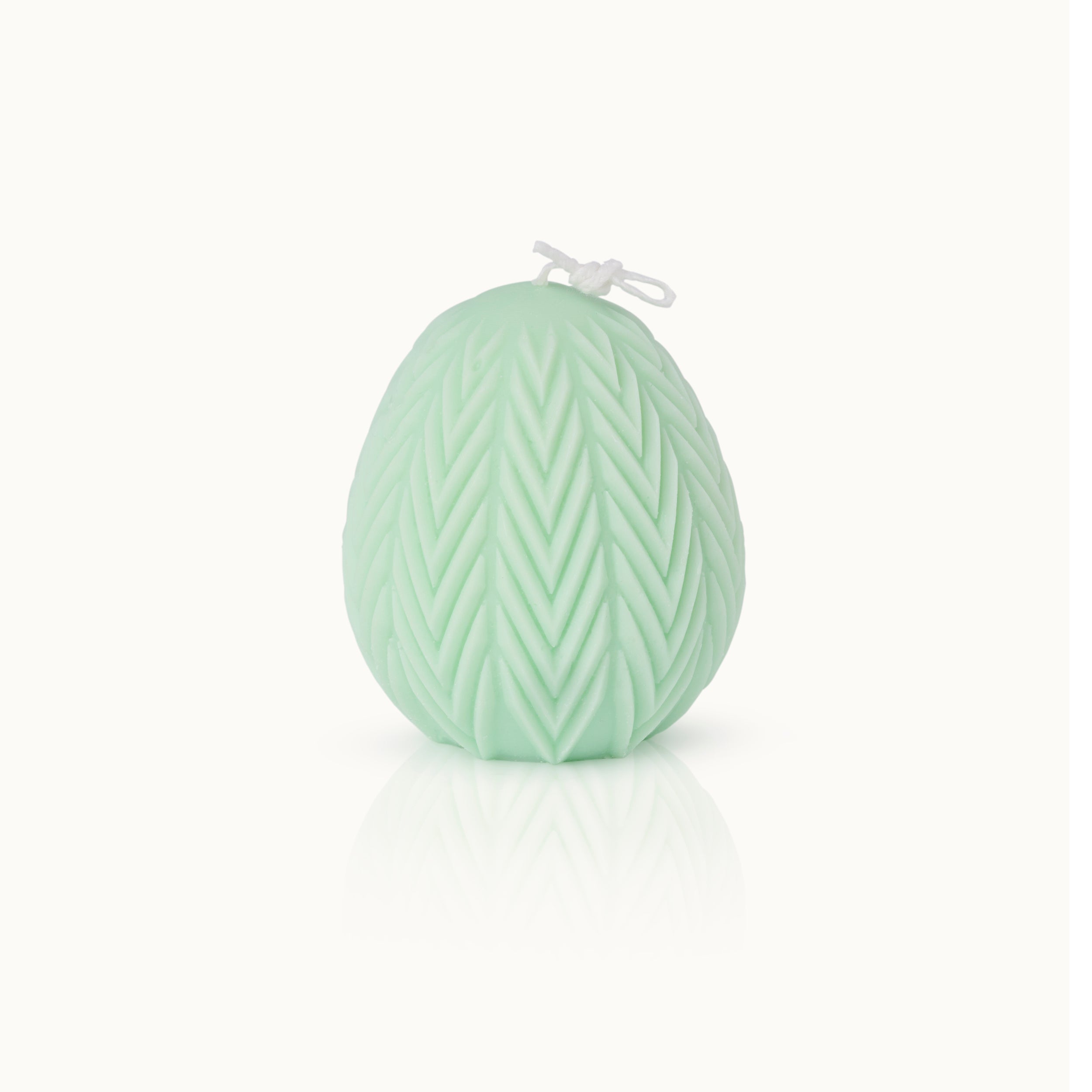 Eine hellgrüne, eiförmige Kerze mit einem strukturierten Chevron-Muster und einem weißen Docht, der oben zusammengebunden ist, vor einem schlichten weißen Hintergrund.