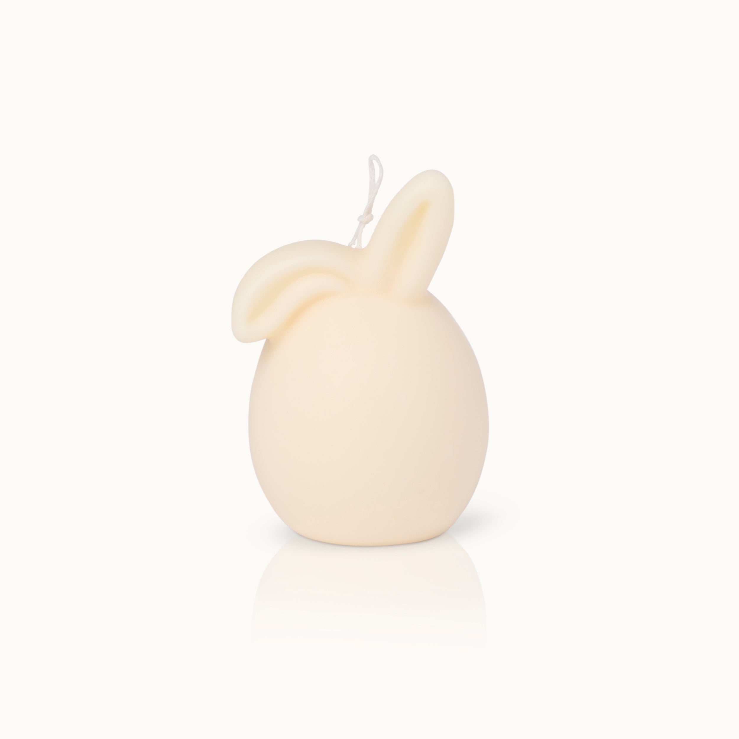 Eine cremefarbene Kerze in Form eines Eies mit zwei Hasenohren auf der Spitze, vor einem schlichten weißen Hintergrund.