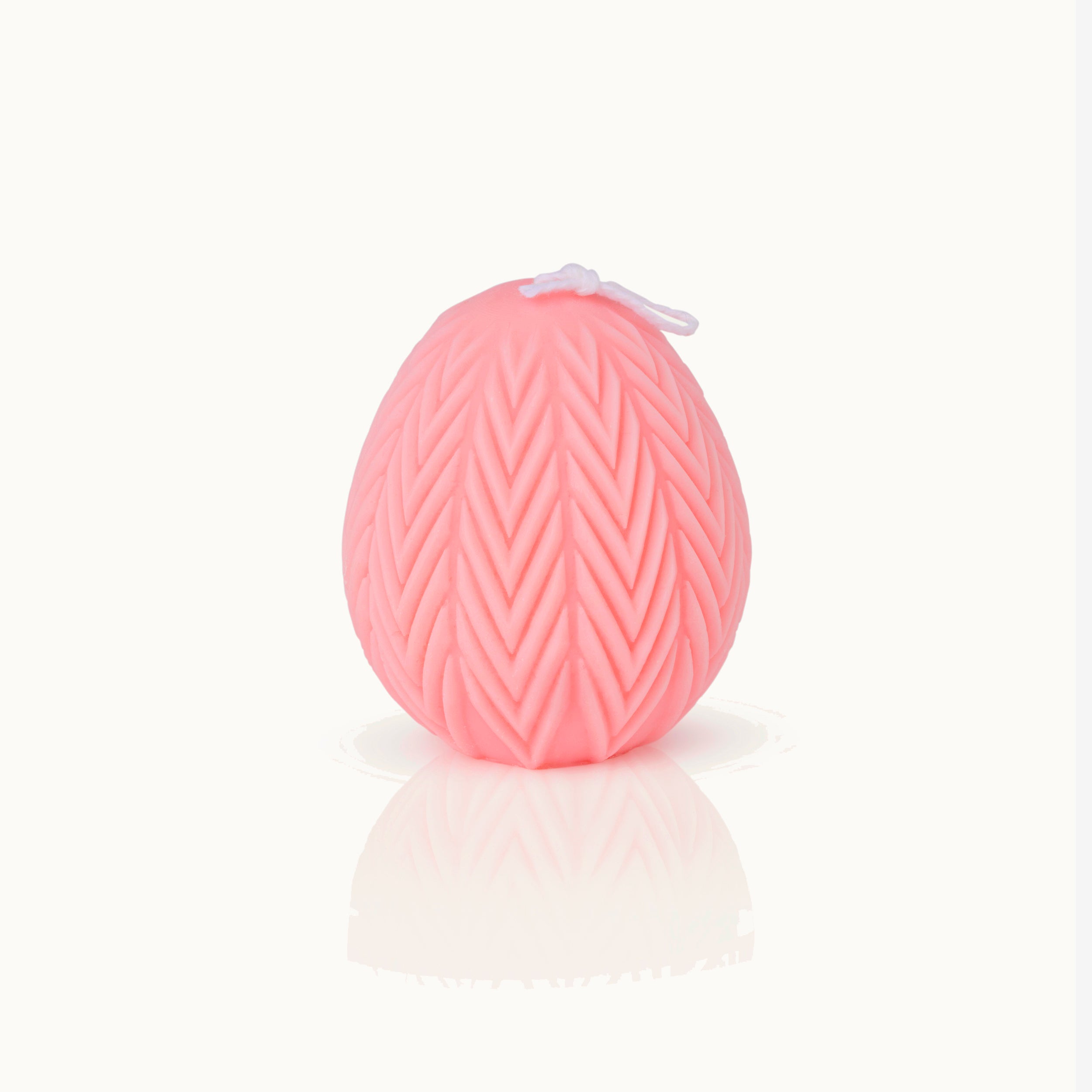Eine rosafarbene, eiförmige Kerze mit einer strukturierten Oberfläche im Chevron-Muster und einem weißen Docht auf der Oberseite, vor einem weißen Hintergrund mit einer schwachen Reflexion darunter.