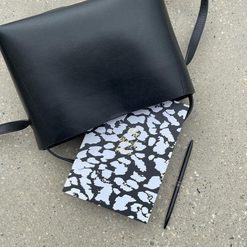 Schwarze Ledertasche, aus der ein Notizbuch mit schwarzem und weißem Muster herausragt, daneben ein schwarzer Stift auf Beton.