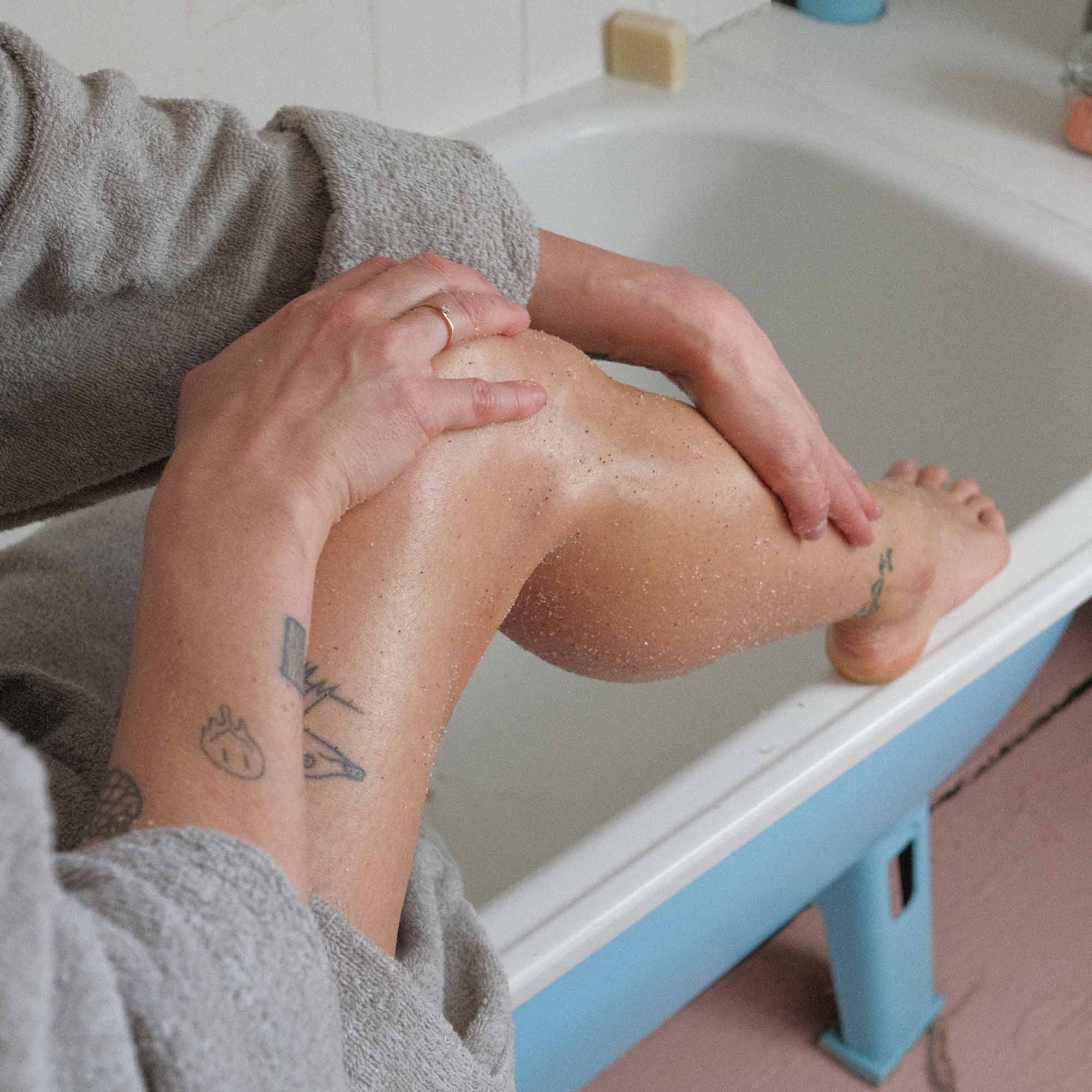Eine Person in einem grauen Bademantel sitzt auf dem Rand einer Badewanne und reibt sich die Beine mit Lotion oder Peeling ein. Ihr Unterarm und ihr Knöchel haben sichtbare Tätowierungen. Das Badezimmer hat eine weiß-blaue Wanne und einen rosa Bodenbelag.
