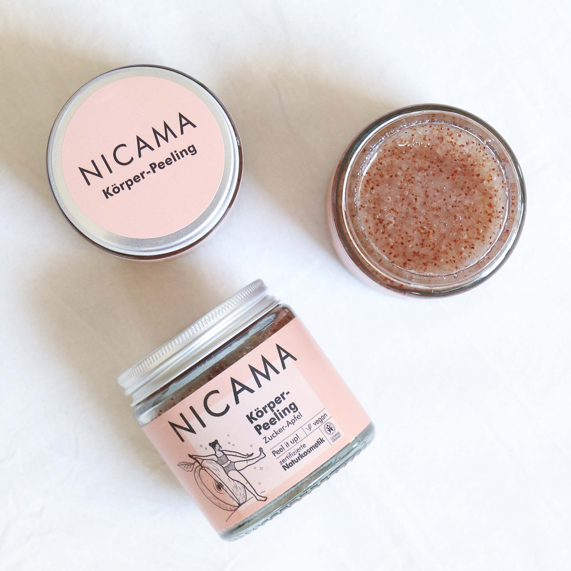 Drei Tiegel des NICAMA Körper-Peelings sind auf einer weißen Fläche ausgestellt; einer ist geschlossen und zeigt das Etikett, einer ist geöffnet und zeigt das Peeling, und einer ist angewinkelt, um das Frontetikett des Produkts zu zeigen.