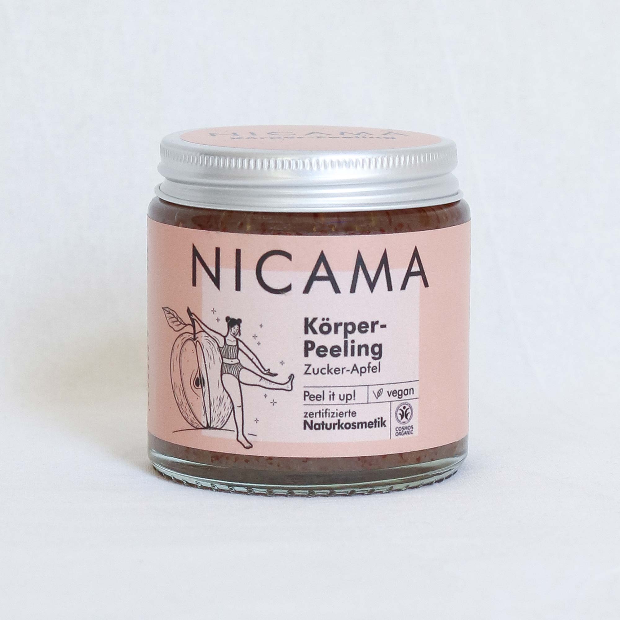 Ein Tiegel NICAMA Körper-Peeling Zucker-Apfel, ein veganes Zucker-Apfel-Körperpeeling. Das Etikett ist rosa und zeigt eine Frau, die einen großen Apfel in der Hand hält, sowie Zertifizierungs- und Vegan-Symbole.