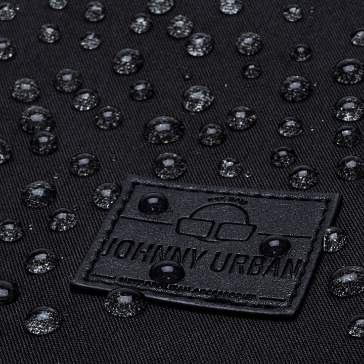 Nahaufnahme von Wassertropfen auf einer schwarzen Stoffoberfläche mit einem rechteckigen Johnny Urban-Aufnäher, der die wasserabweisende Eigenschaft des Materials demonstriert.