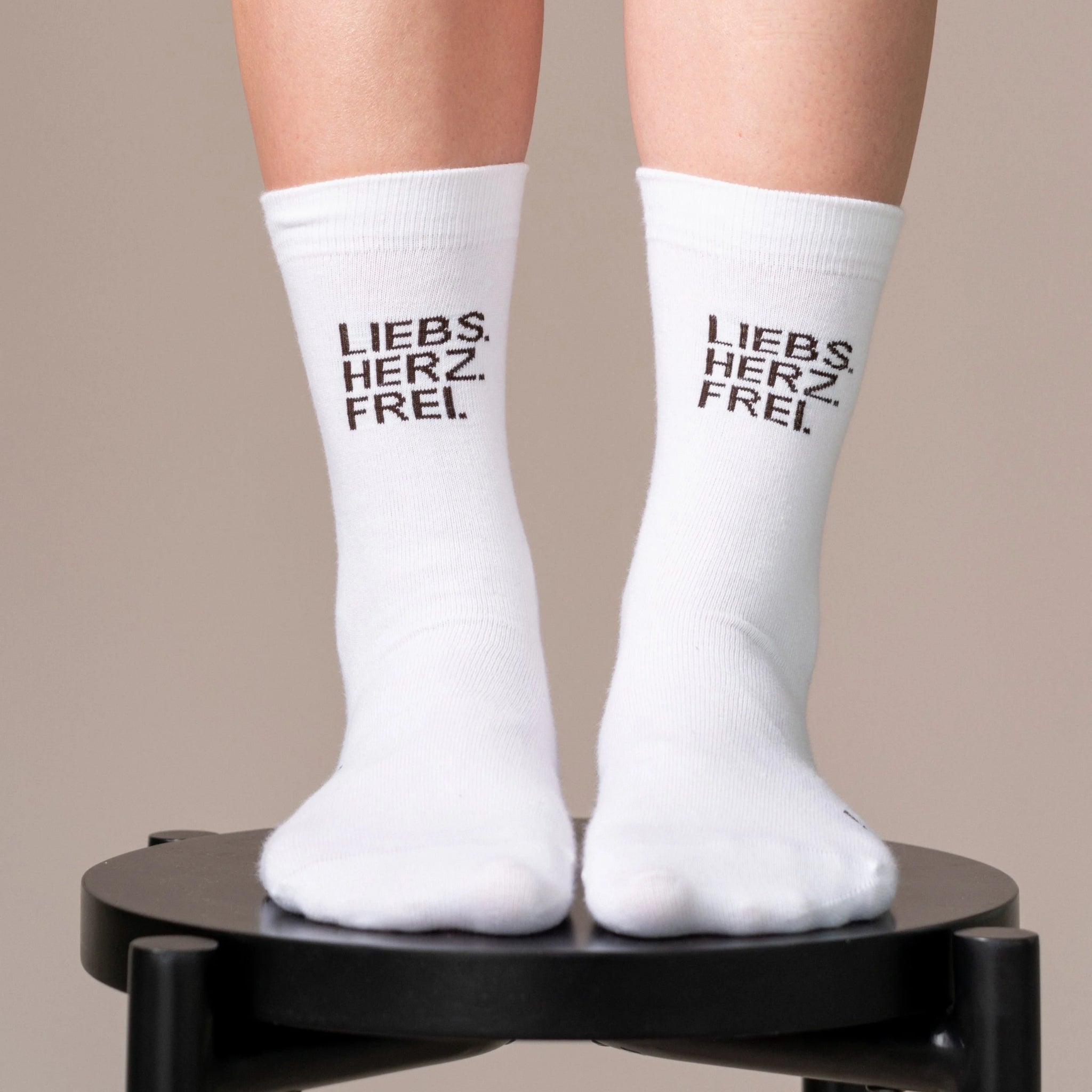 Ein Paar Füße in weißen Socken mit der Aufschrift LIEBS. HERZ. FREI. in schwarzer Schrift auf einem schwarzen Hocker vor einem neutralen Hintergrund stehend.