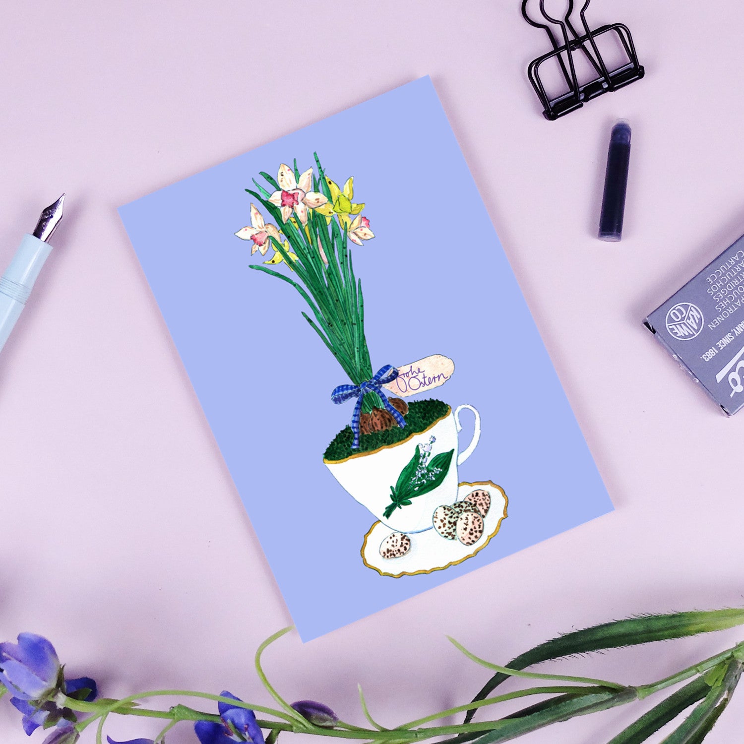Eine Grußkarte mit einer Illustration von Narzissen und anderen Blumen, die aus einer Teetasse, Erde und einem Ei mit der Aufschrift Frohe Ostern wachsen. Die Karte ruht auf einer hellvioletten Oberfläche mit einem Stift, einem Clip und Blumen in der Nähe.