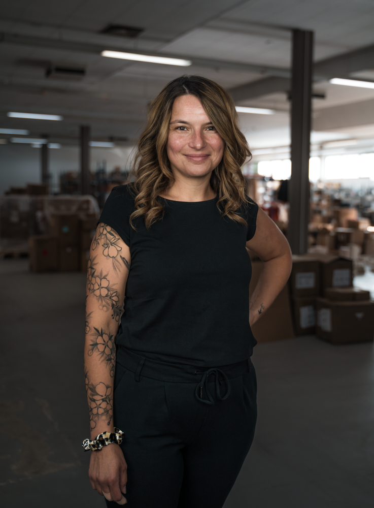 Eine Frau steht in einem Lagerraum, lächelt in die Kamera und trägt ein schwarzes Outfit mit Blumen-Tattoo am Arm.