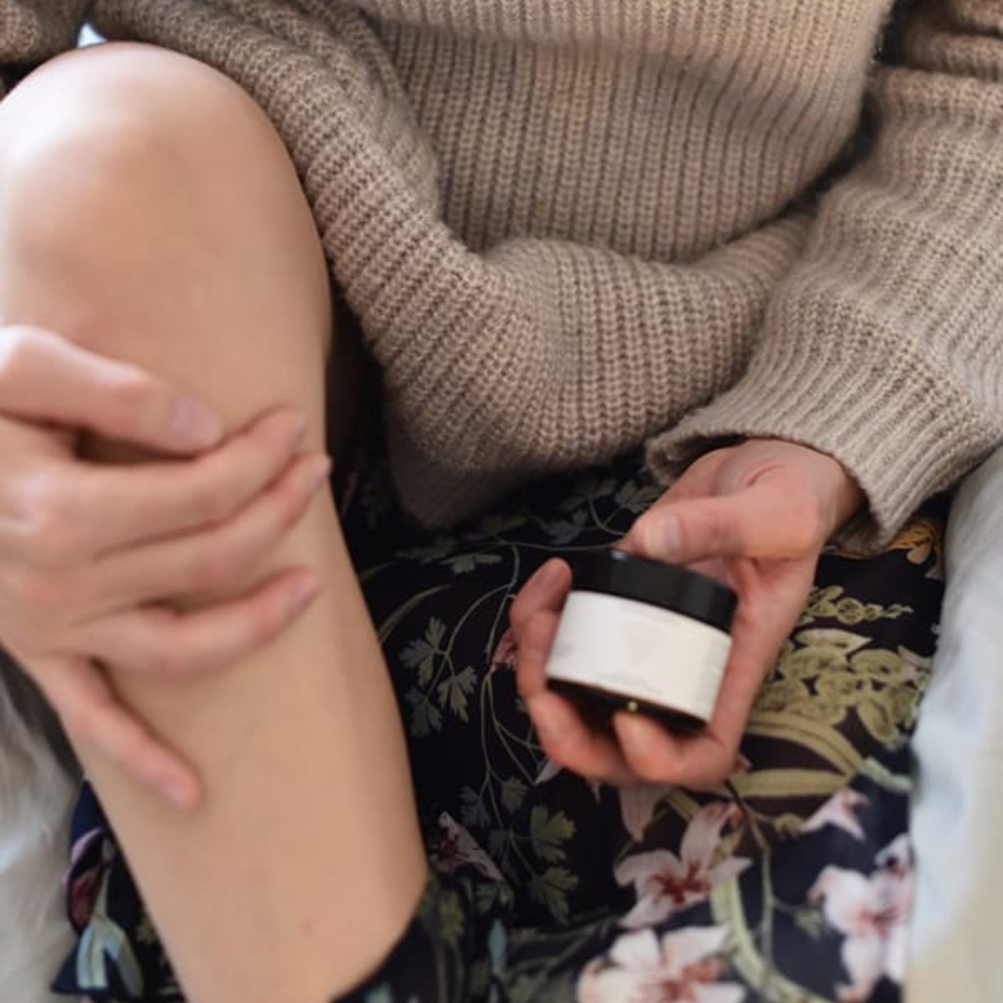 Person in wool sweater holding cream jar, applying it to bare leg.