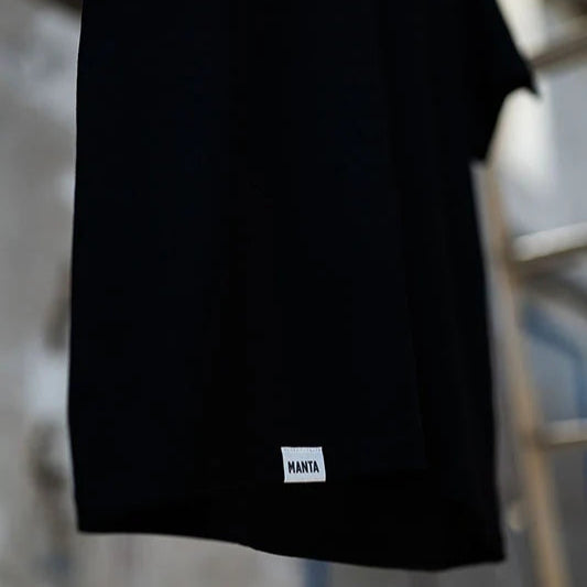 Schwarzes T-Shirt mit dem Etikett "MANTA" am Saum vor unscharfem Hintergrund.