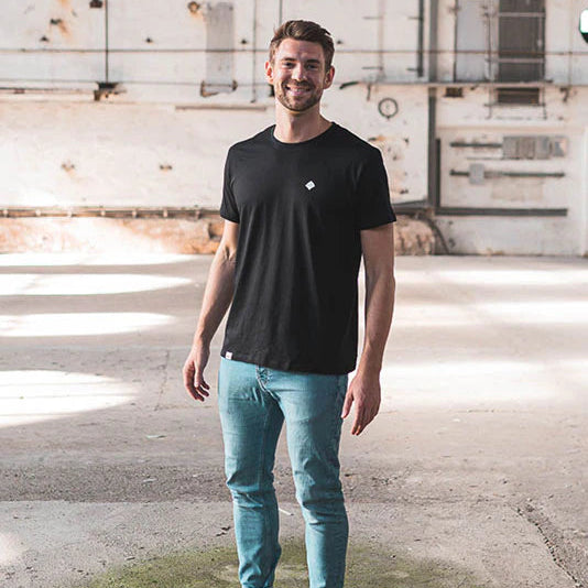 Ein Mann in einem schwarzen T-Shirt und Jeans steht lächelnd in einem hellen, leeren Industriegebäude.