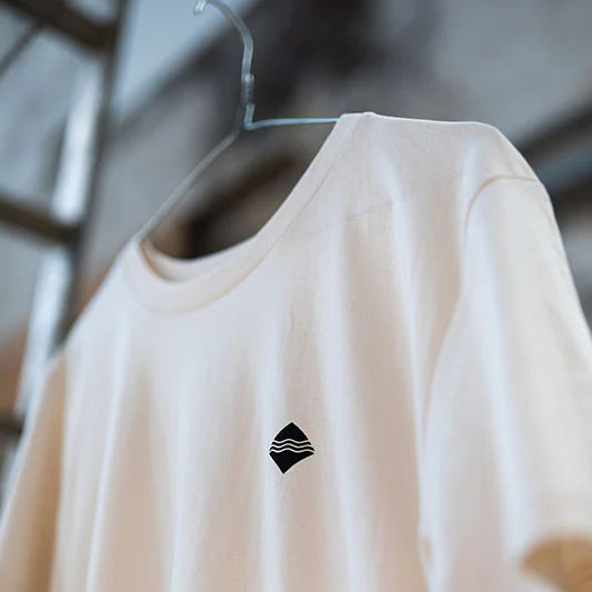 Ein beigefarbenes T-Shirt mit einem kleinen schwarzen Symbol hängt an einem Kleiderbügel vor einem unscharfen Hintergrund.