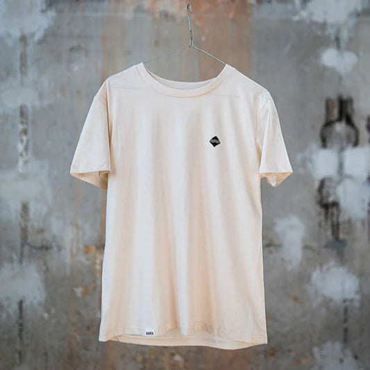 Beiges T-Shirt mit kleinem Logo hängt an einem Kleiderbügel vor einer gemusterten, verwitterten Betonwand.
