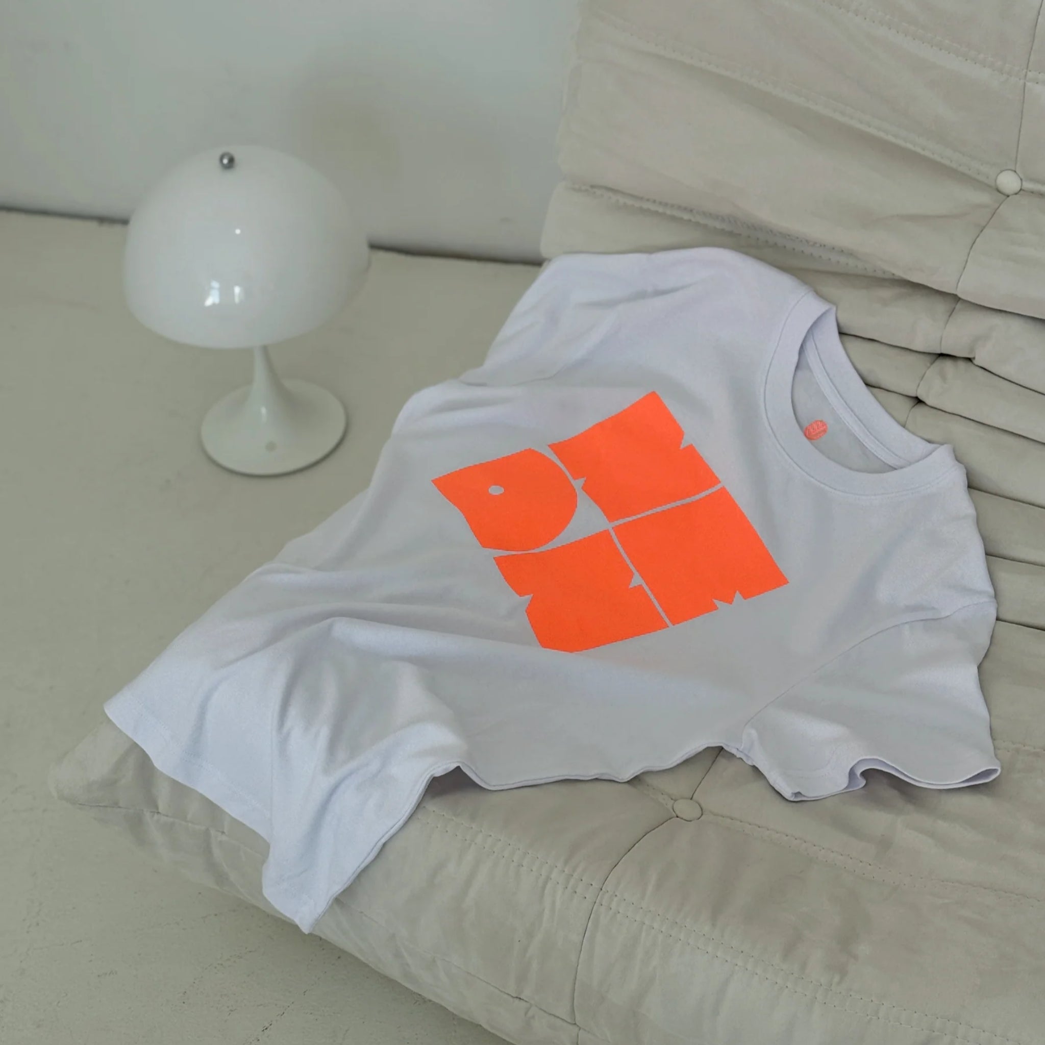 Ein weißes T-Shirt mit einer abstrakten, orangefarbenen Grafik liegt auf einem cremefarbenen, gepolsterten Stuhl neben einer kleinen, modernen weißen Lampe.