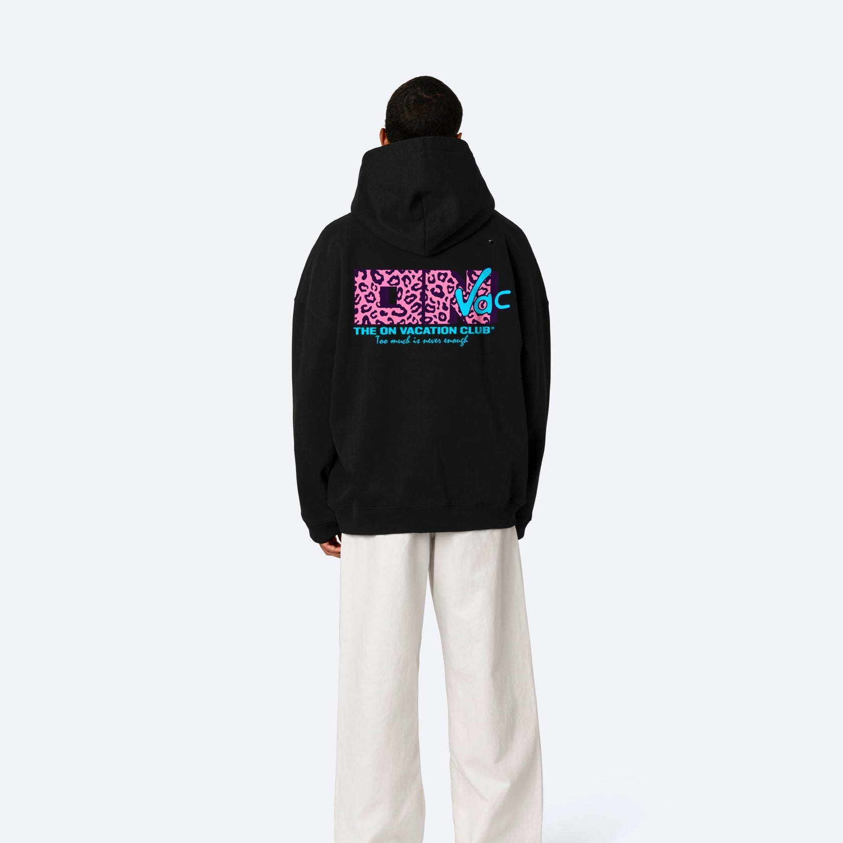 Eine Person steht mit dem Gesicht zur Wand, trägt einen schwarzen Kapuzenpulli mit hellrosa und blauer Grafik auf dem Rücken und eine weite weiße Hose vor einem schlichten hellgrauen Hintergrund.