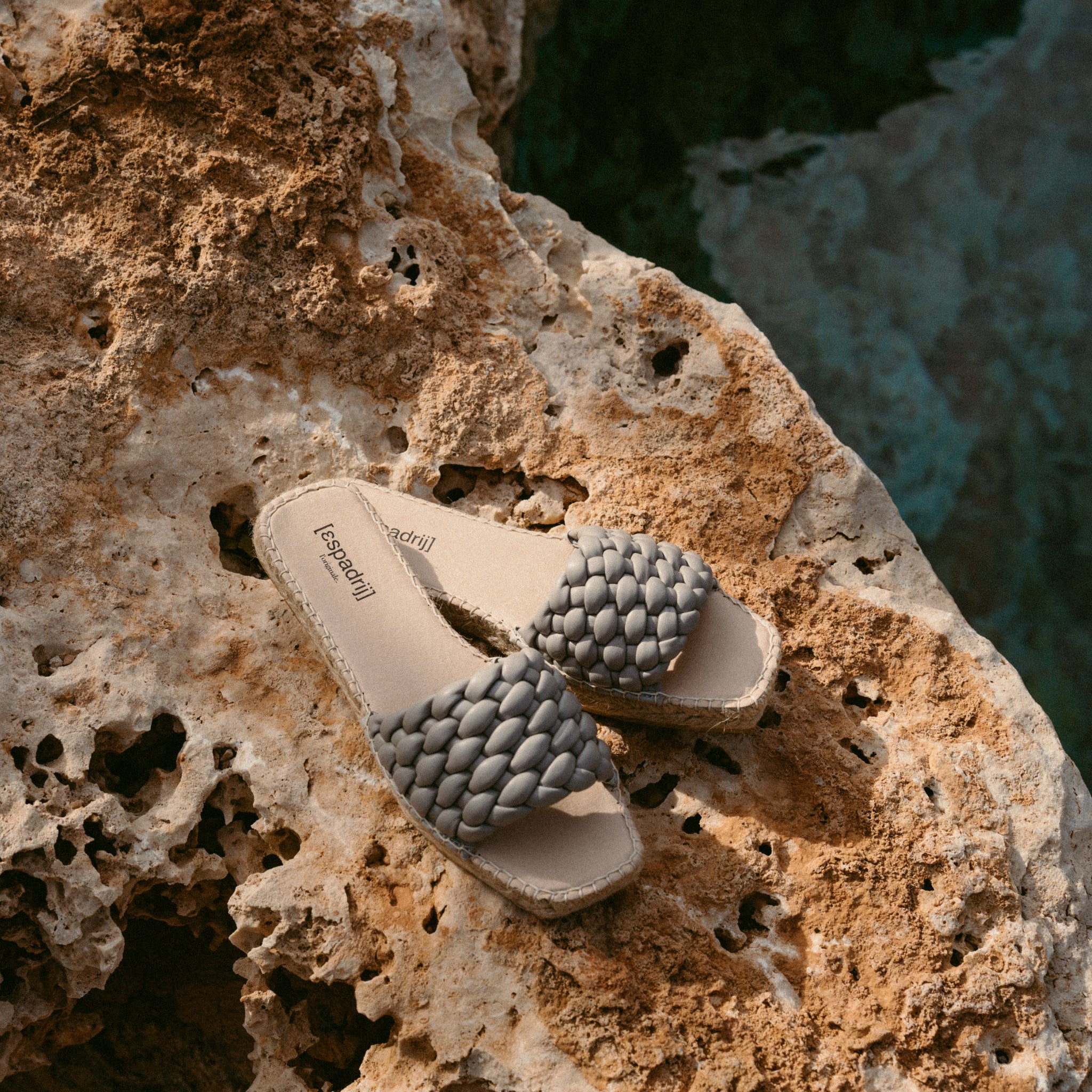 Beige Sandalen mit geflochtenem Riemen liegen auf einem rauen, felsigen Untergrund.