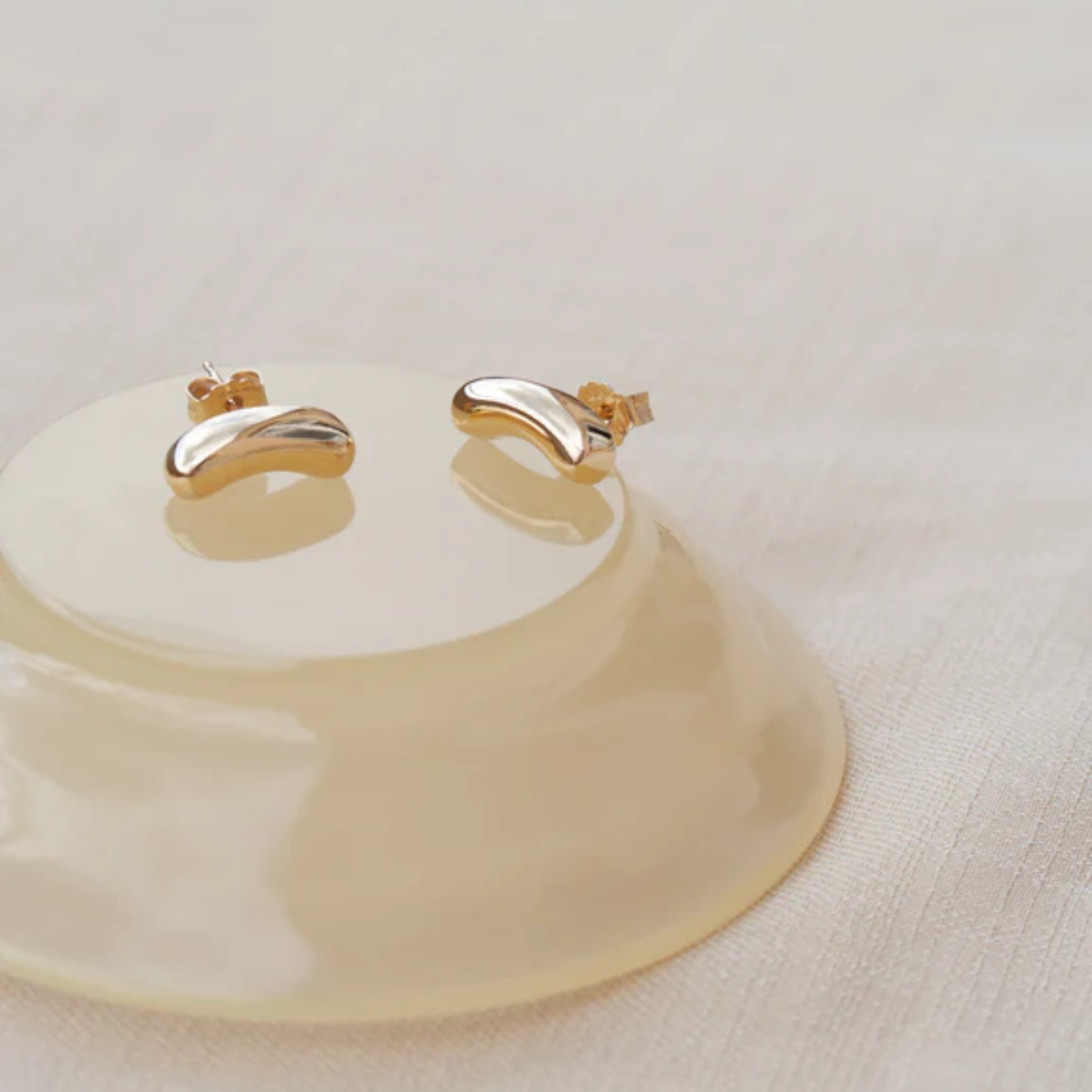 Kleine goldene Ohrringe in Bohnenform auf einer cremefarbenen, glänzenden Oberfläche liegend.