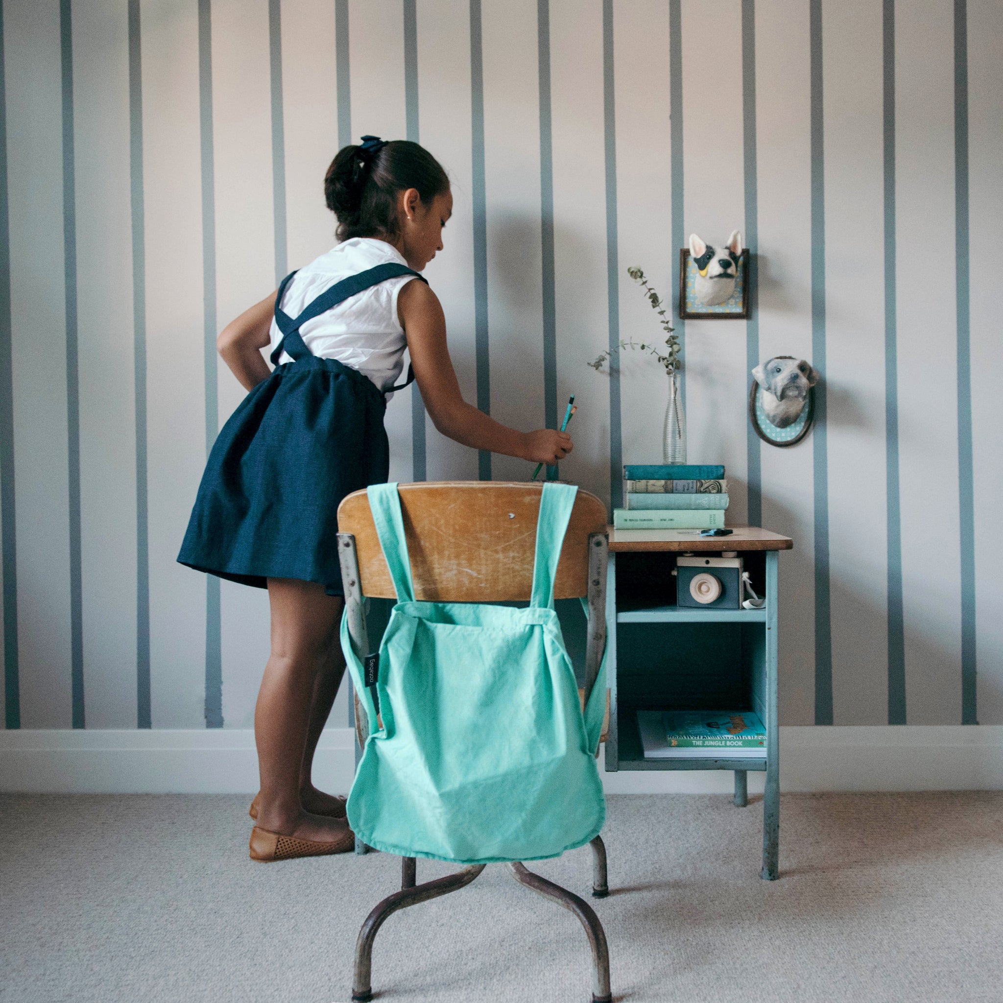 Mädchen in Schuluniform steht an einem kleinen Schreibtisch mit Büchern und Schreibzeug, Rucksack auf dem Stuhl.