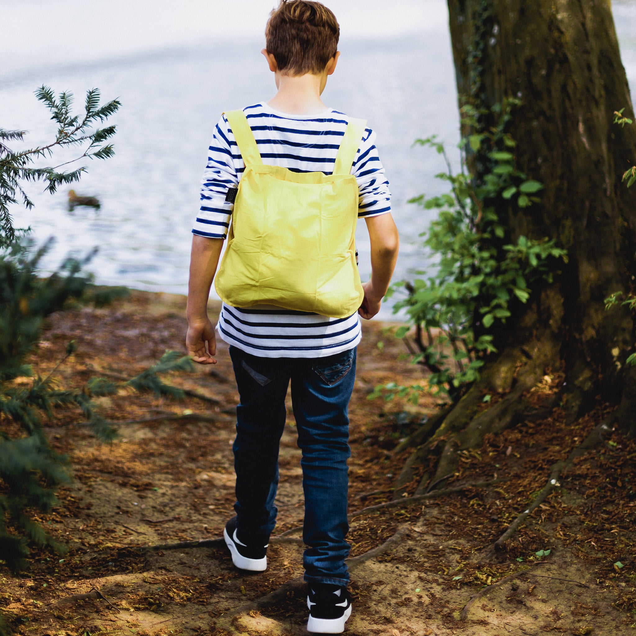 Junge mit gestreiftem Shirt und gelbem Rucksack geht einen Waldweg entlang, nahe einem Gewässer.